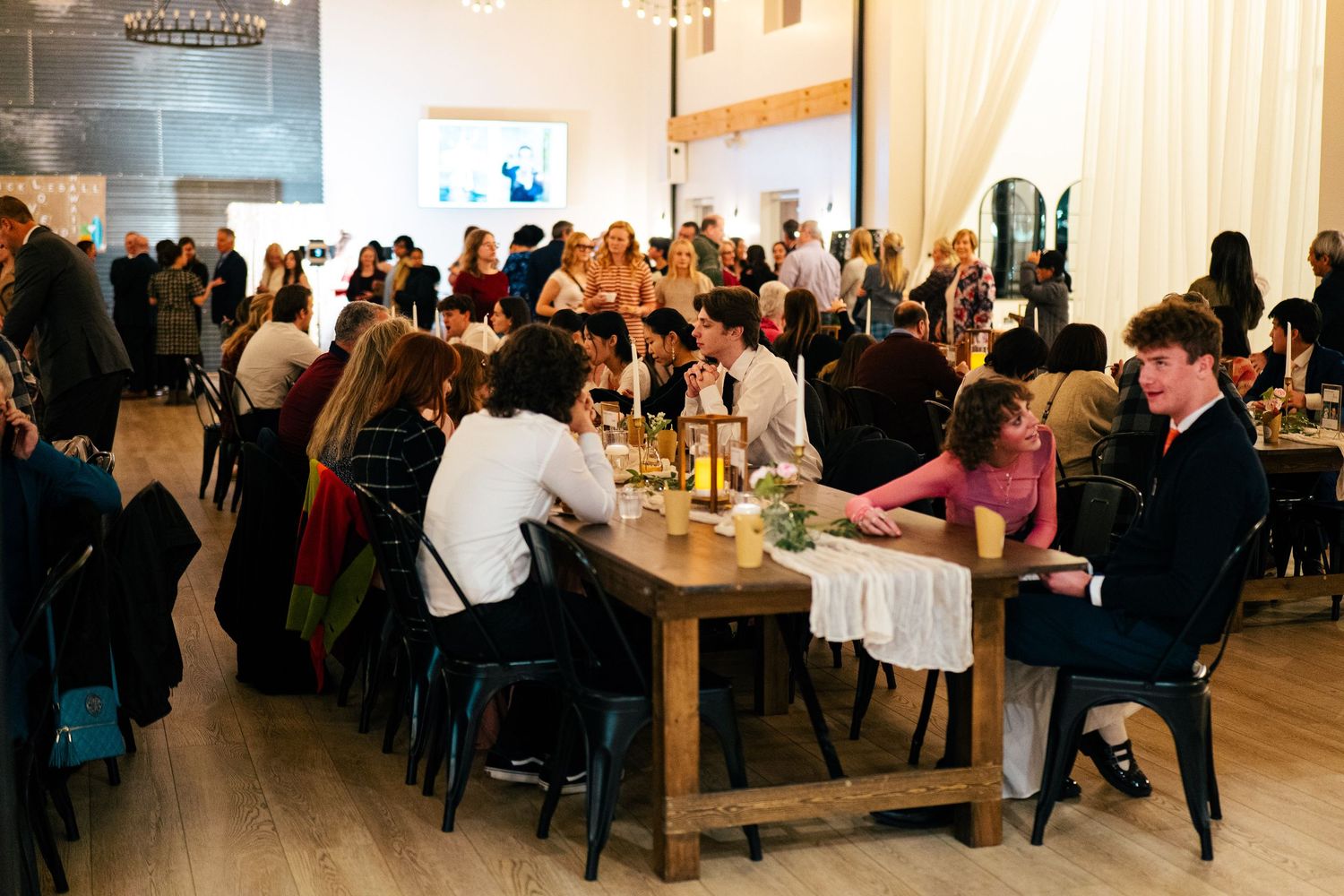 Guests dining at long wooden tables in an elegant event venue with white curtains and warm lighting.