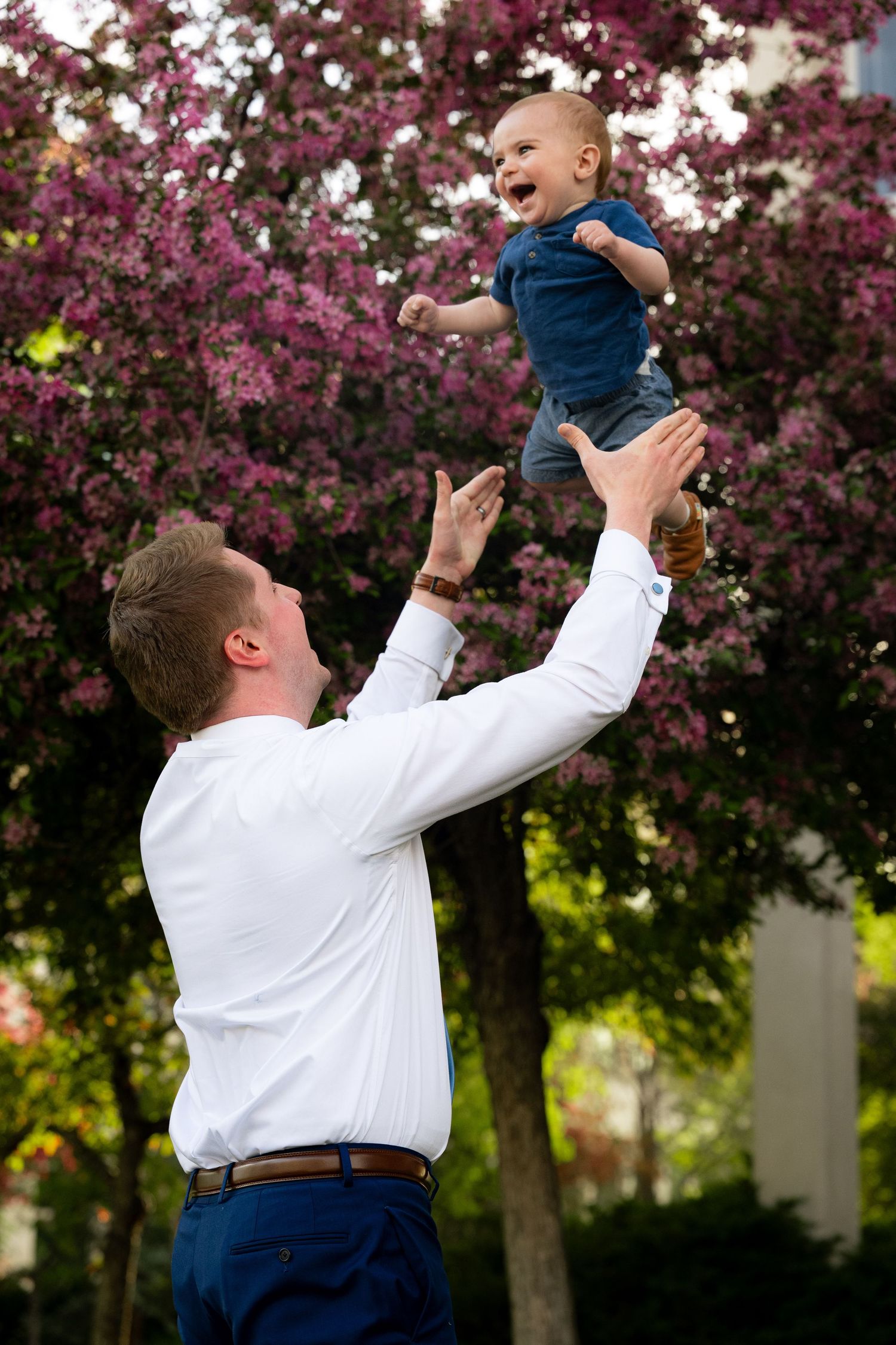 A man is joyfully lifting up a smiling baby in a garden with blooming flowers.