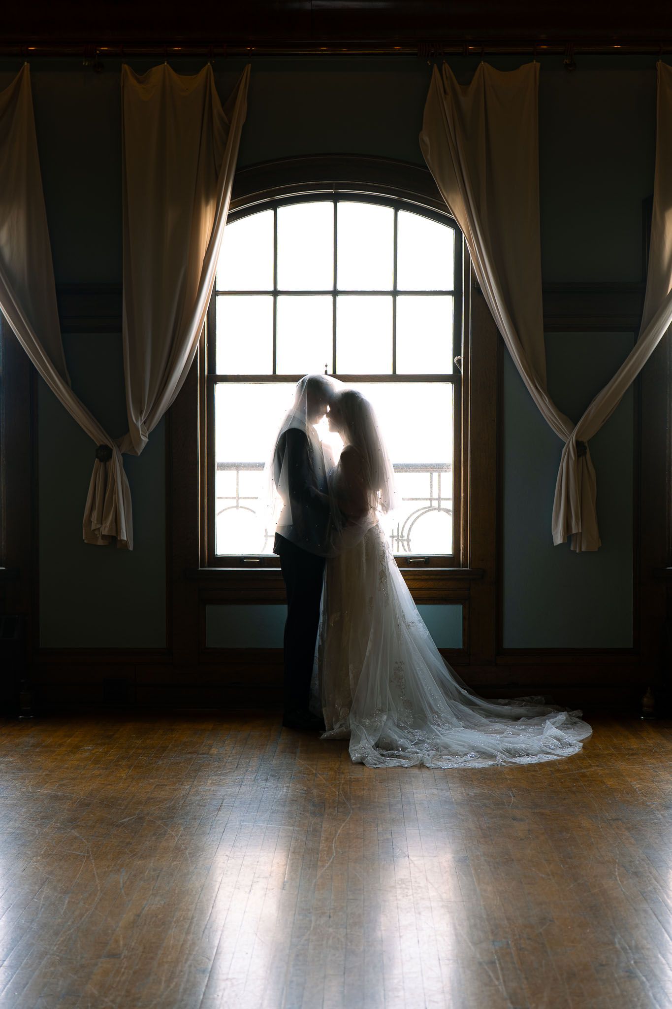 Bride and groom silhouetted against arched window with flowing curtains on hardwood floor.