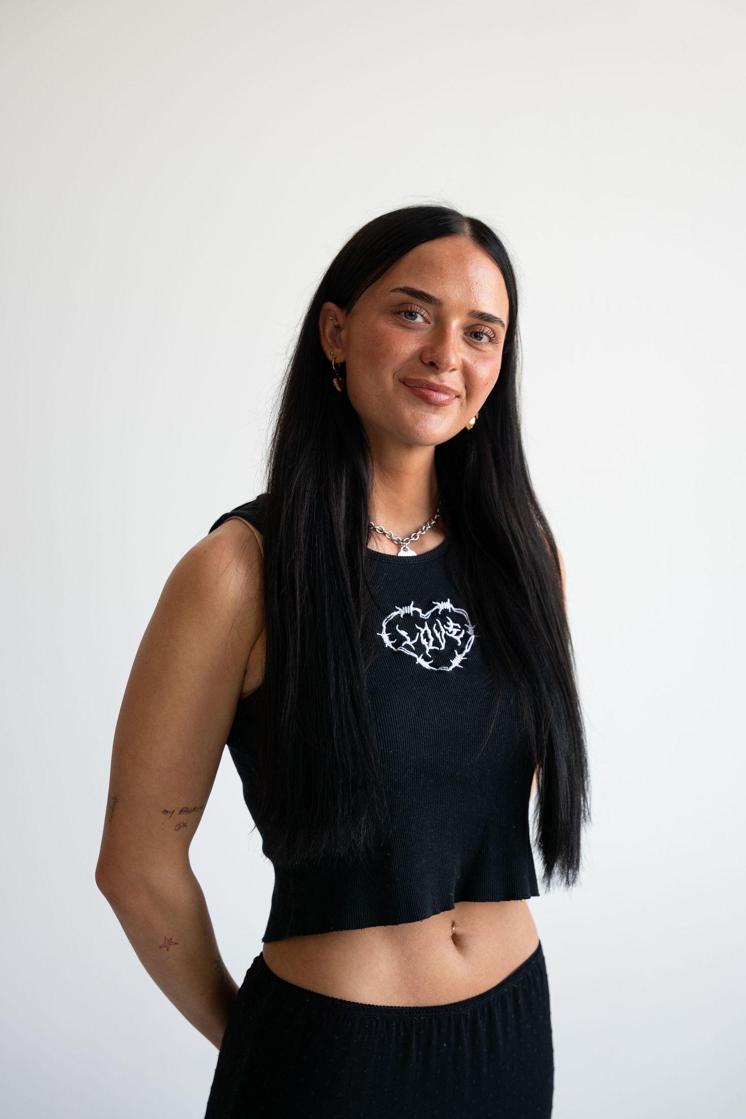 Young woman with long dark hair wearing a black crop top with graphic print in a white studio.