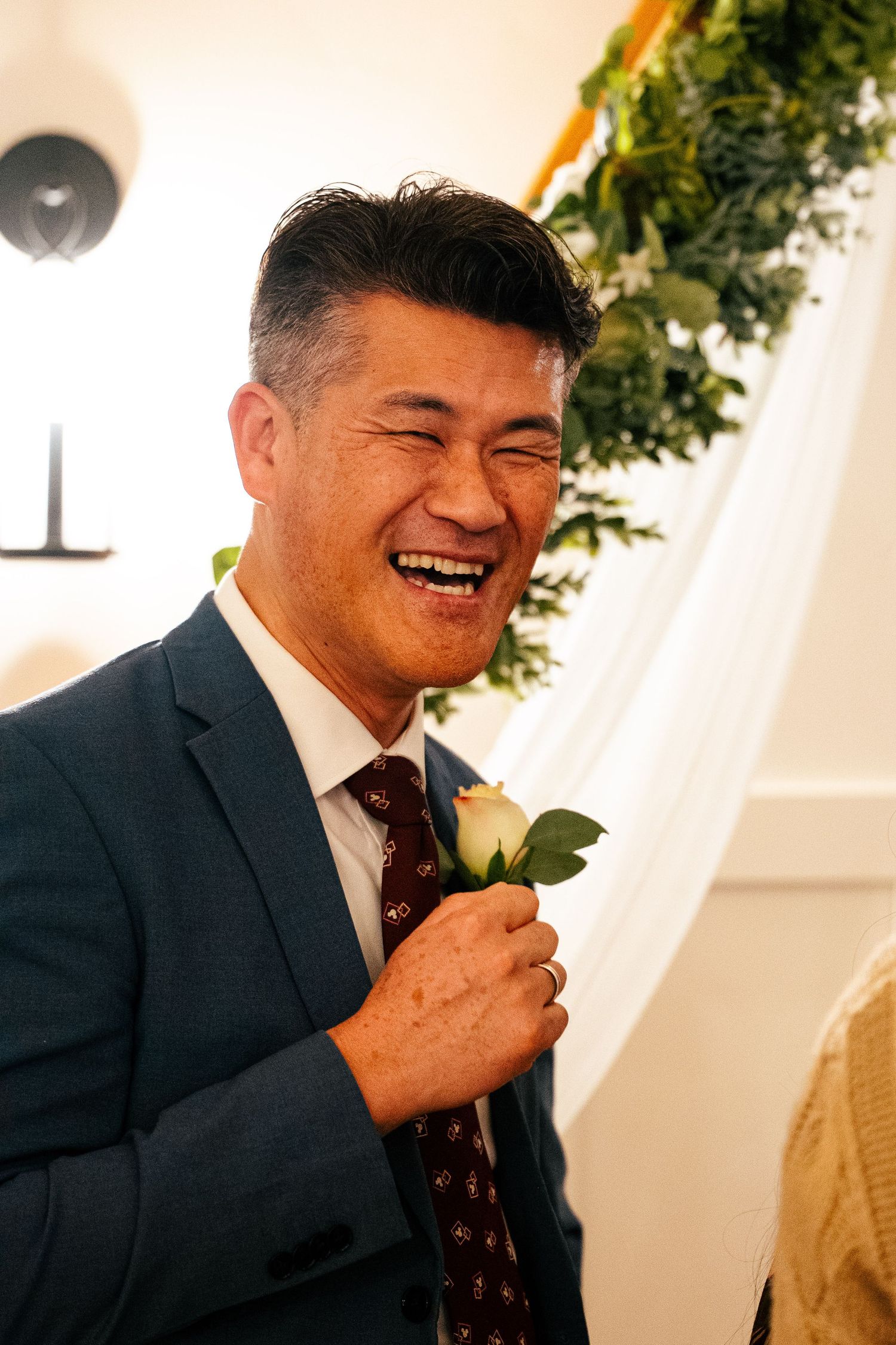 Man in navy suit laughing joyfully while touching boutonniere at wedding ceremony.