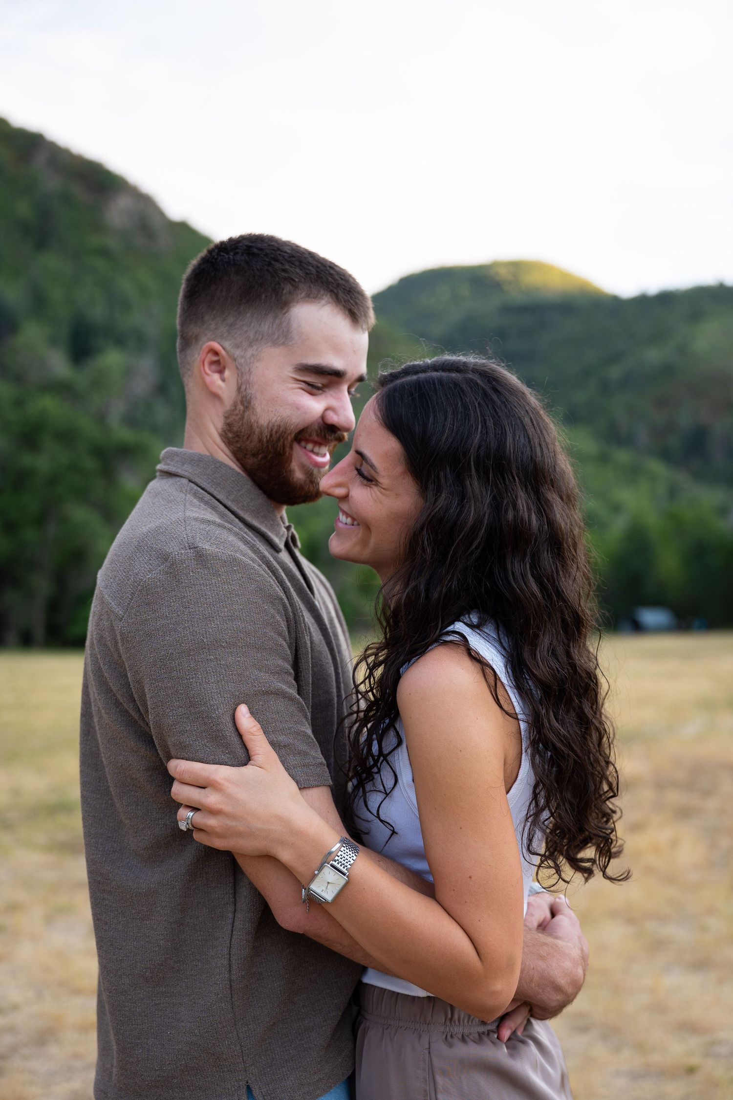 A smiling couple embraces in a mountain meadow, sharing a romantic moment with green hills in the background.