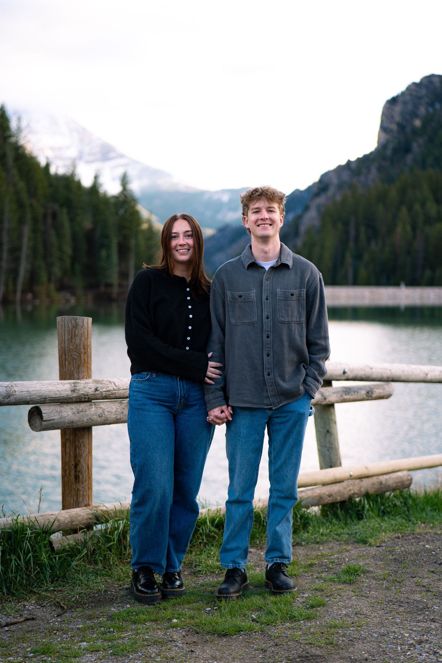 A young couple holds hands smiling by a rustic log fence with a mountain lake and forest backdrop.