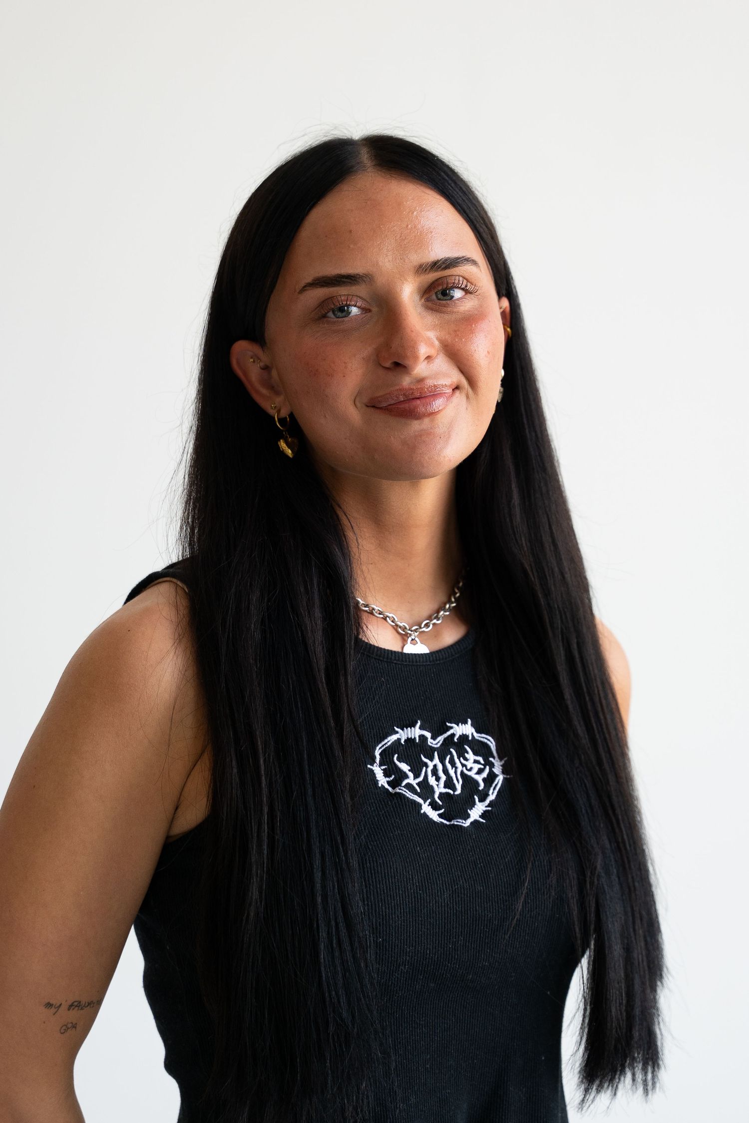 Young woman with long black hair wearing black graphic tank top and chain necklace against white background.