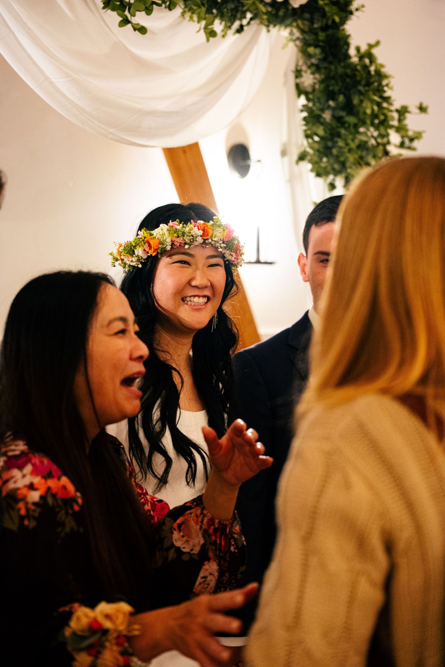 Joyful bride wearing colorful flower crown laughs with wedding guests during reception celebration.