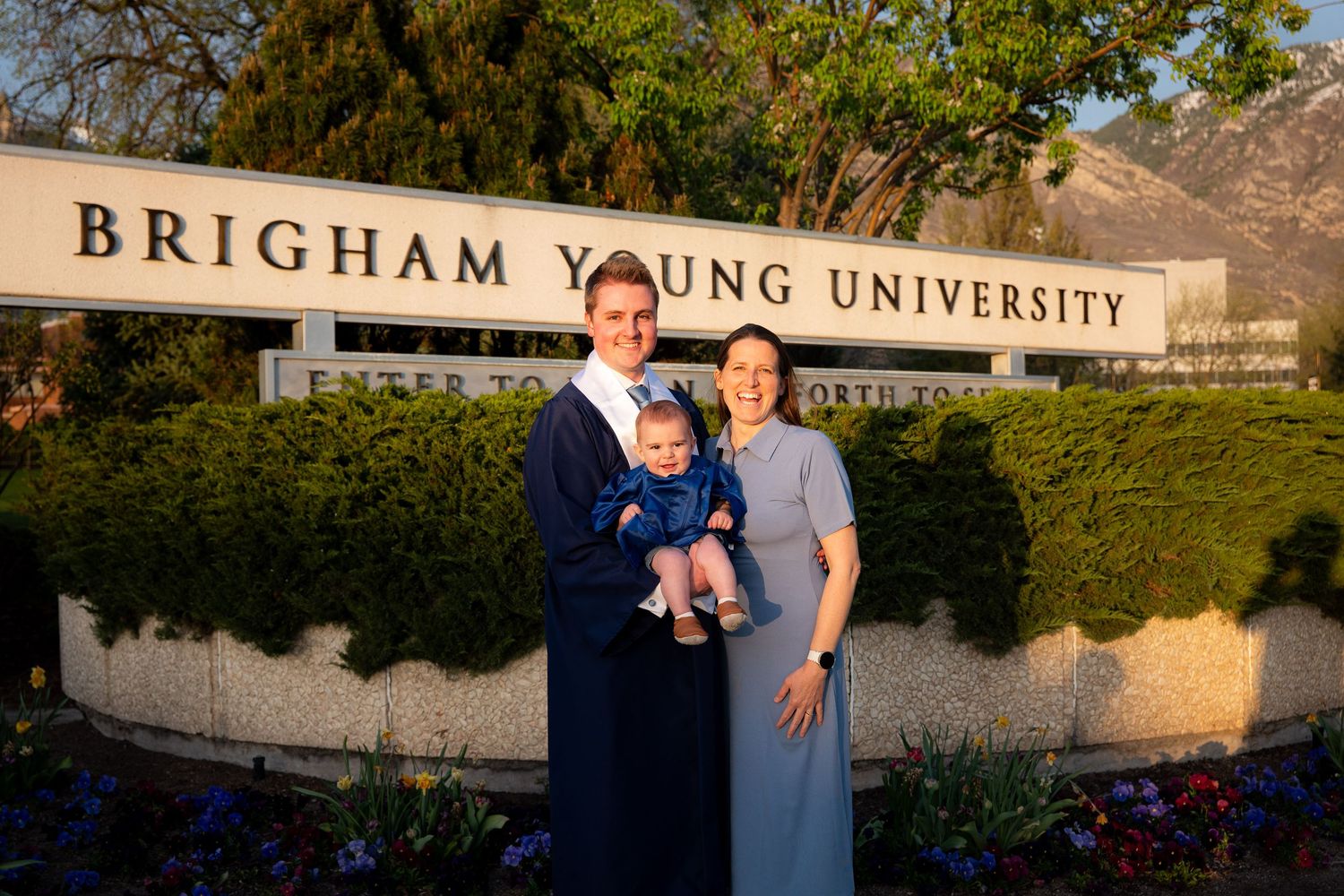 A family of three, including a young child, stands in front of a sign for Brigham Young University.