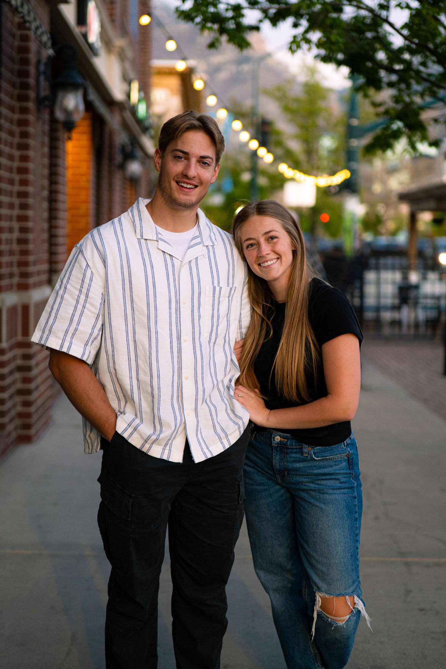 A smiling young couple poses together on a charming brick sidewalk with string lights in the background.