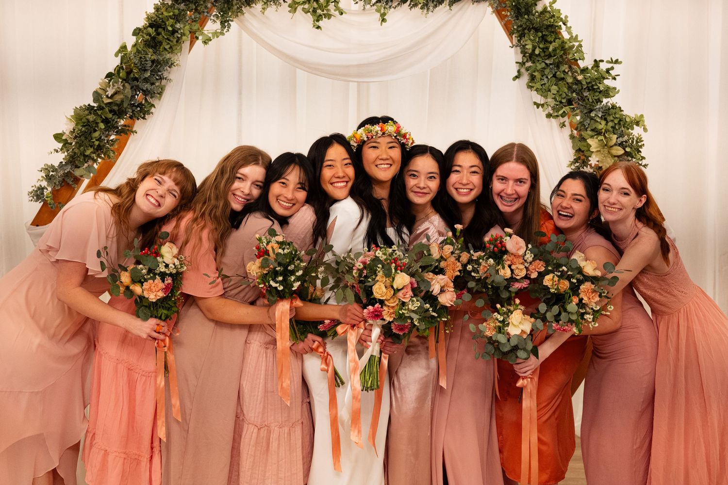 Bride in white gown poses with eight bridesmaids in blush pink dresses holding colorful bouquets under floral arch.