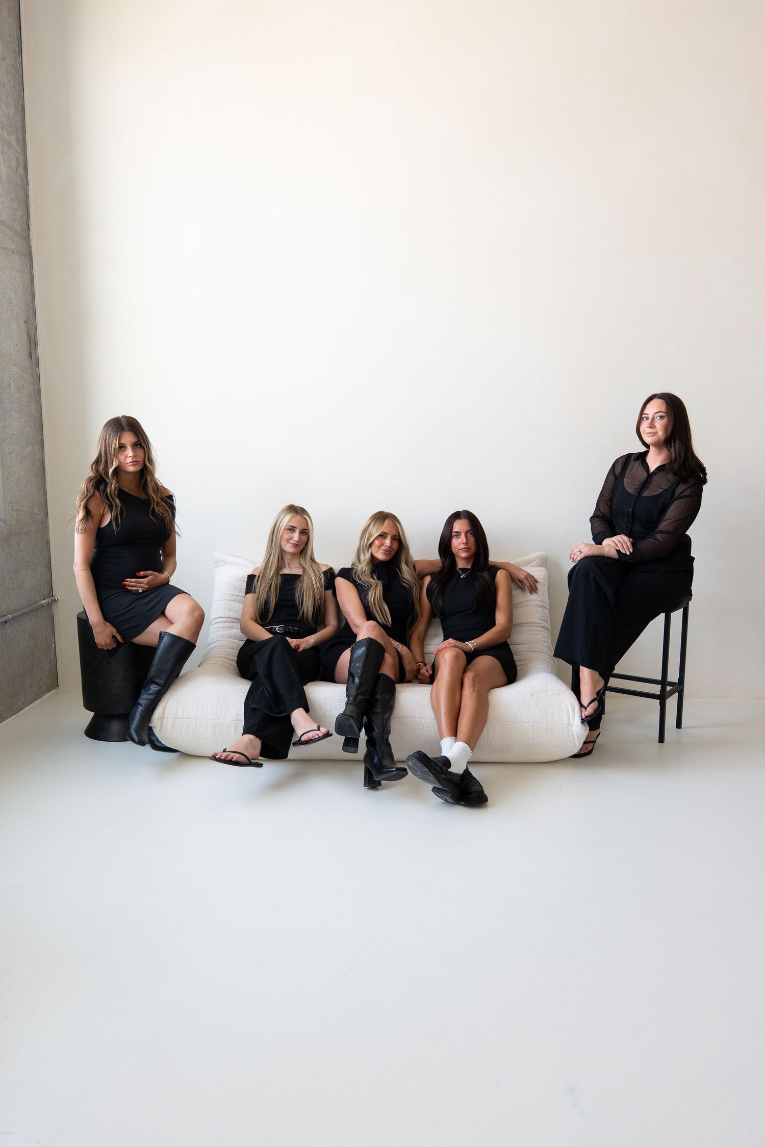 Five women in black outfits pose on a white couch in a minimalist studio setting.