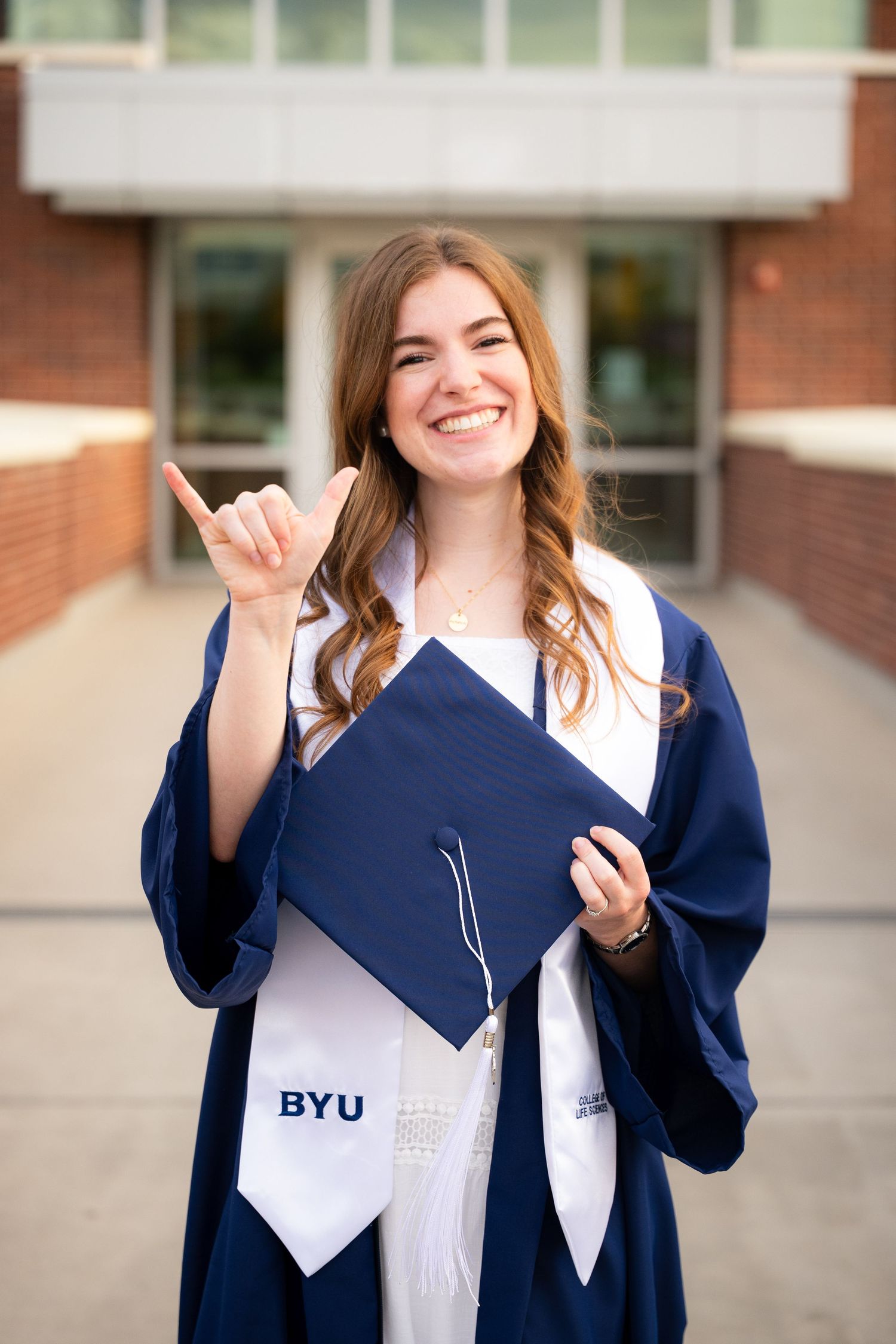 A smiling woman in a graduation gown and cap stands in front of a university building.