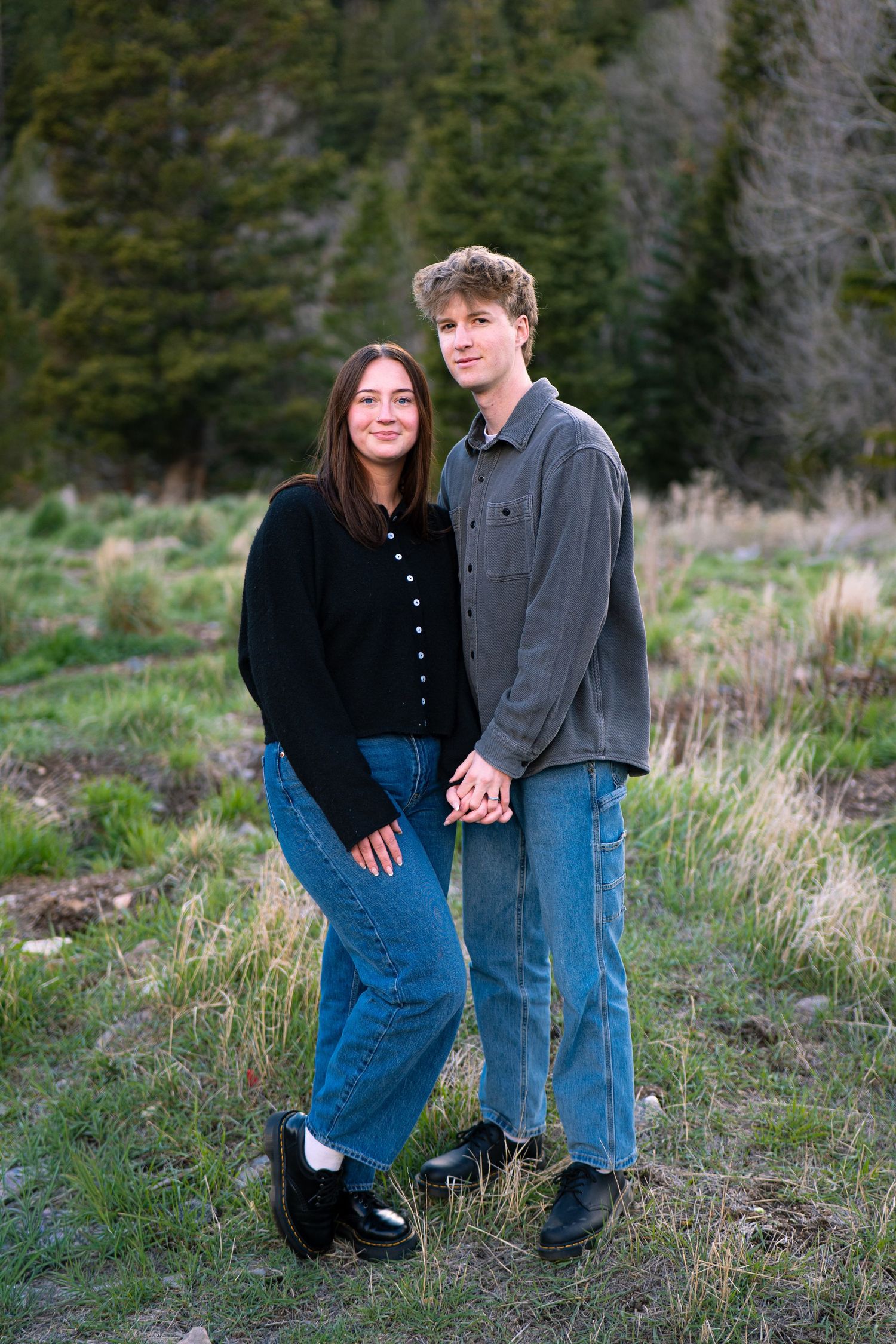Young couple holding hands in a scenic mountain meadow, wearing casual jeans and dark tops, smiling at the camera.
