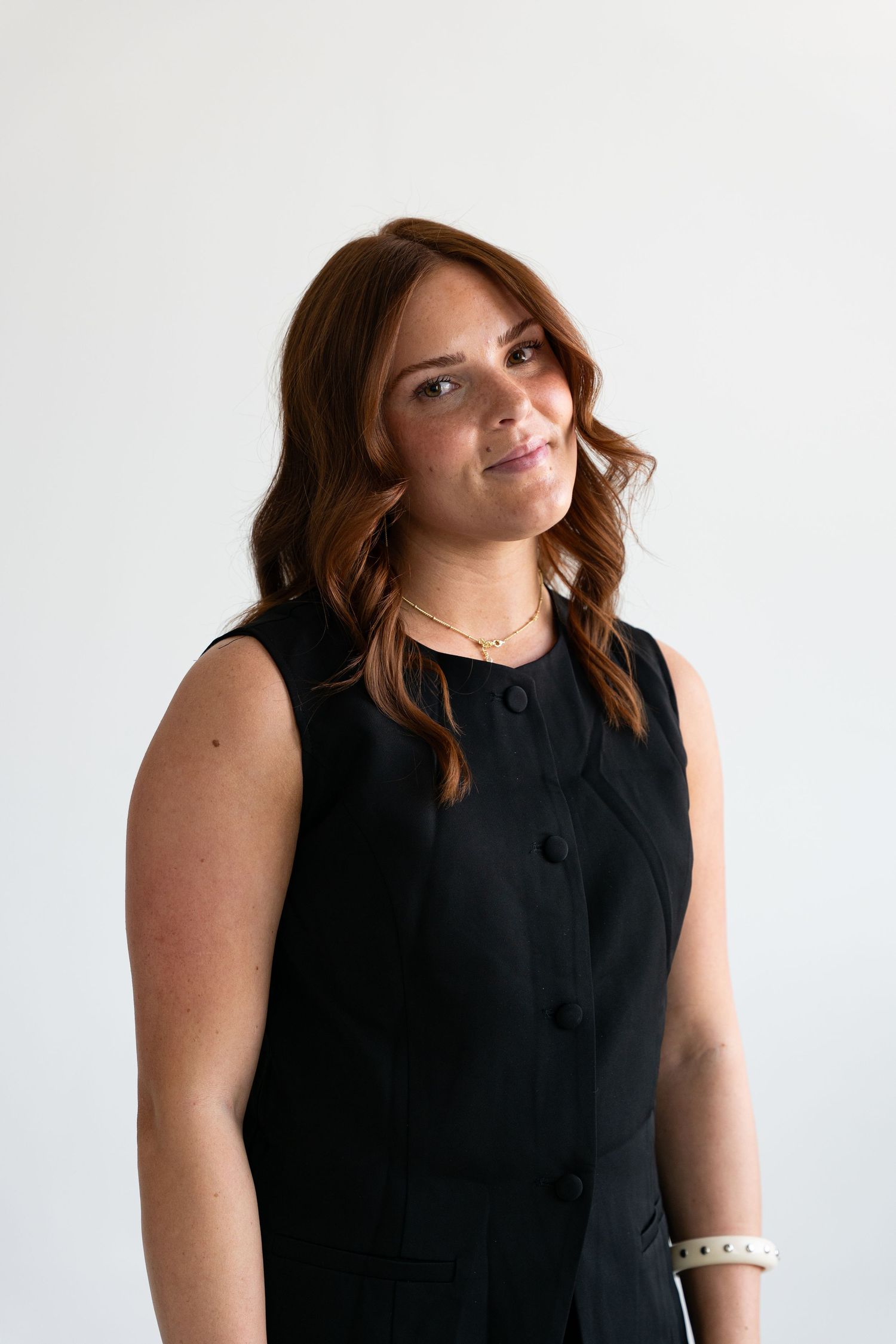 Young woman with auburn hair wearing a black sleeveless dress and gold necklace, smiling against a white background.