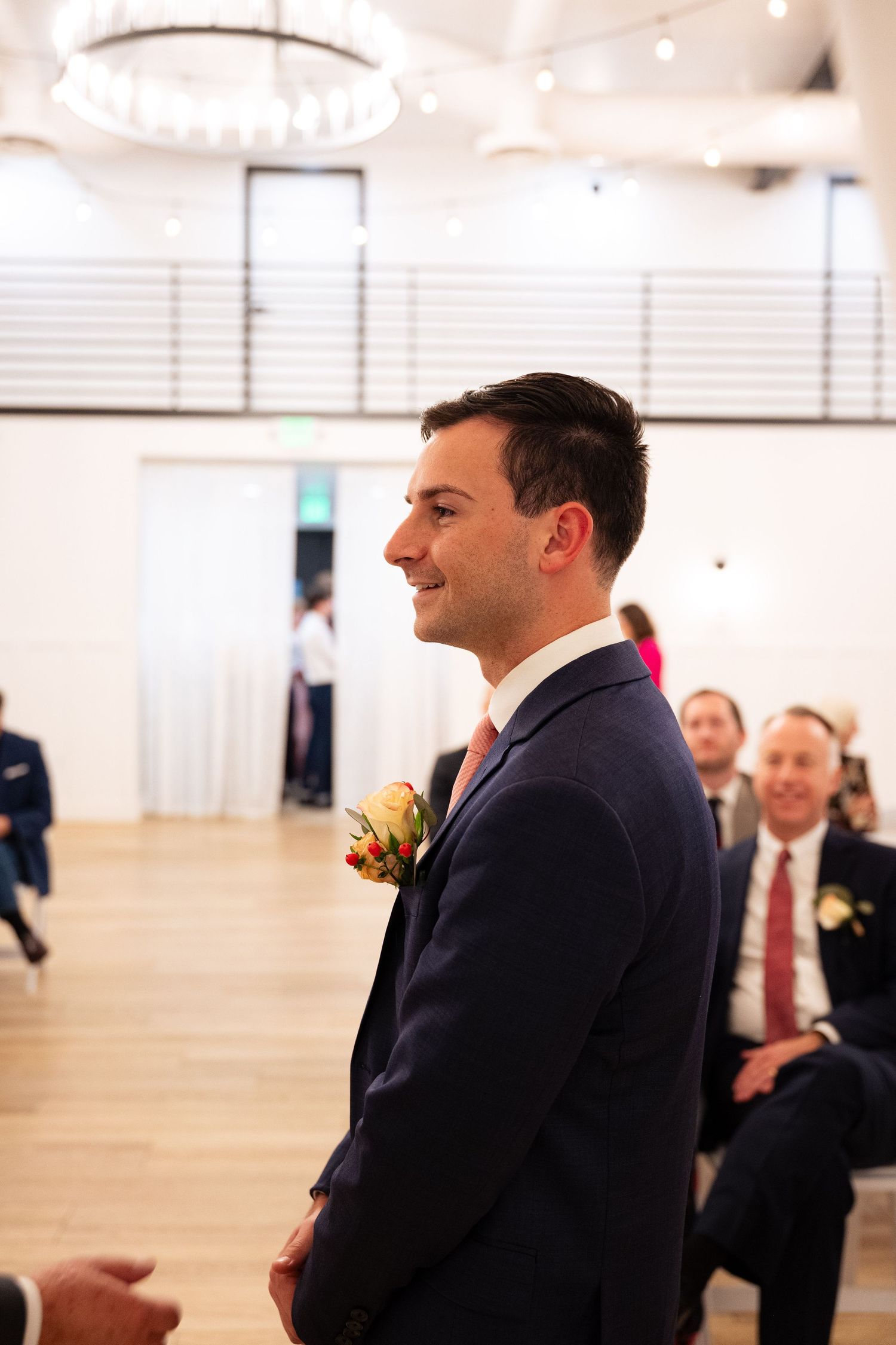 Groom in navy suit with floral boutonniere smiling at wedding ceremony aisle.