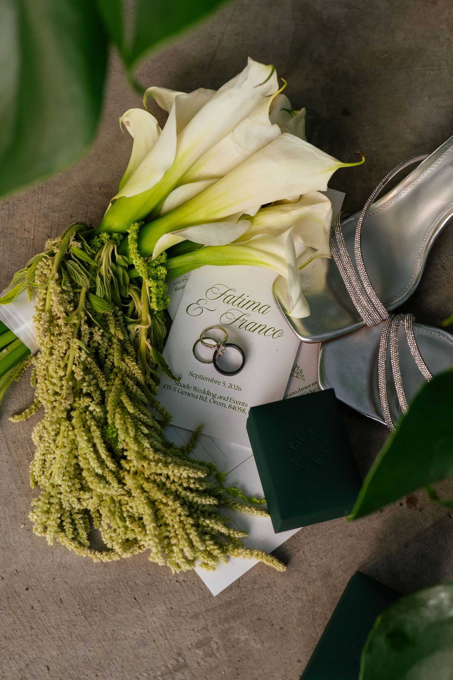 Wedding flat lay with white calla lilies, green amaranthus, silver heels, ring box, and elegant invitation cards on linen.
