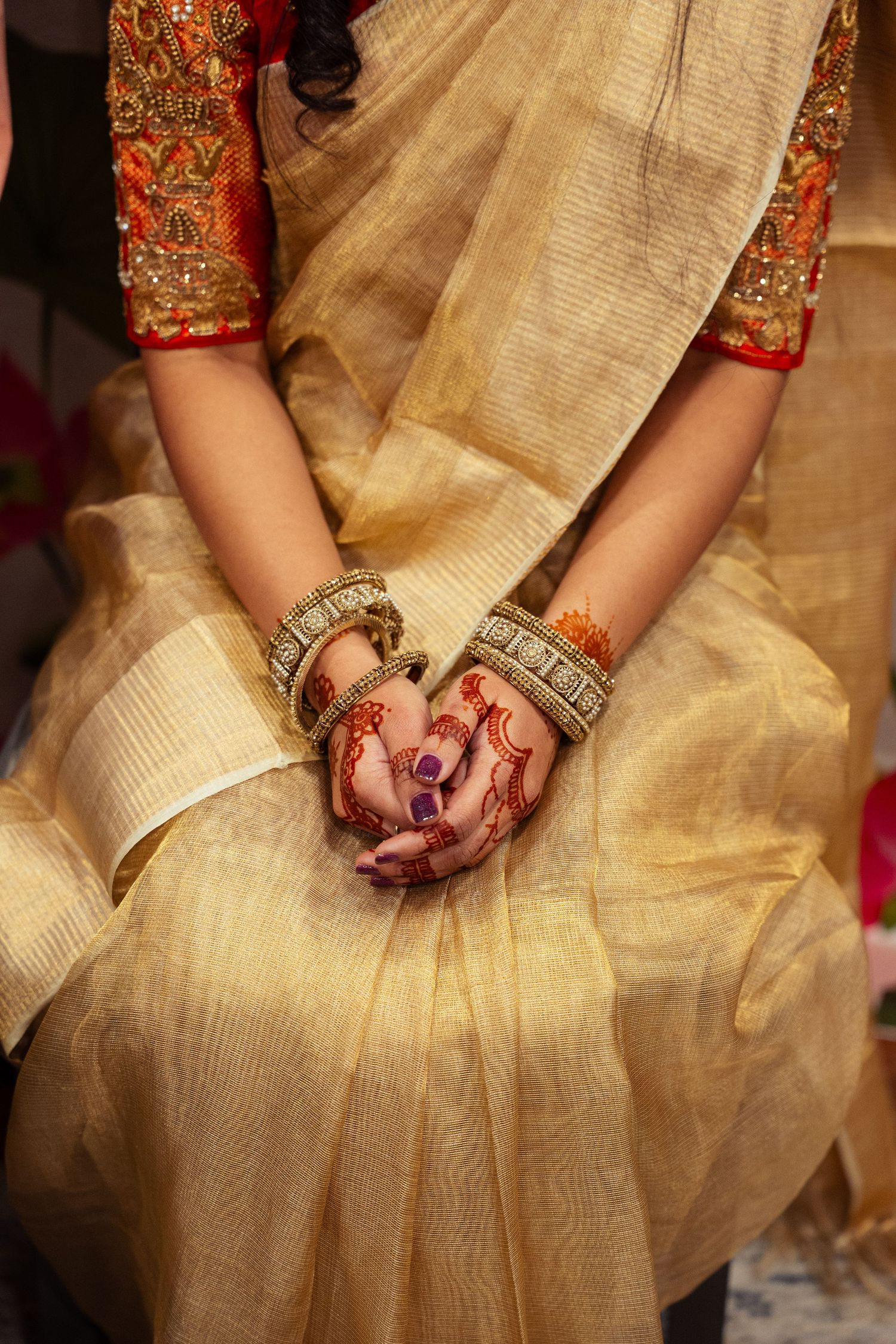 Indian bride in gold saree with henna-adorned hands and traditional bangles resting gracefully on her lap.