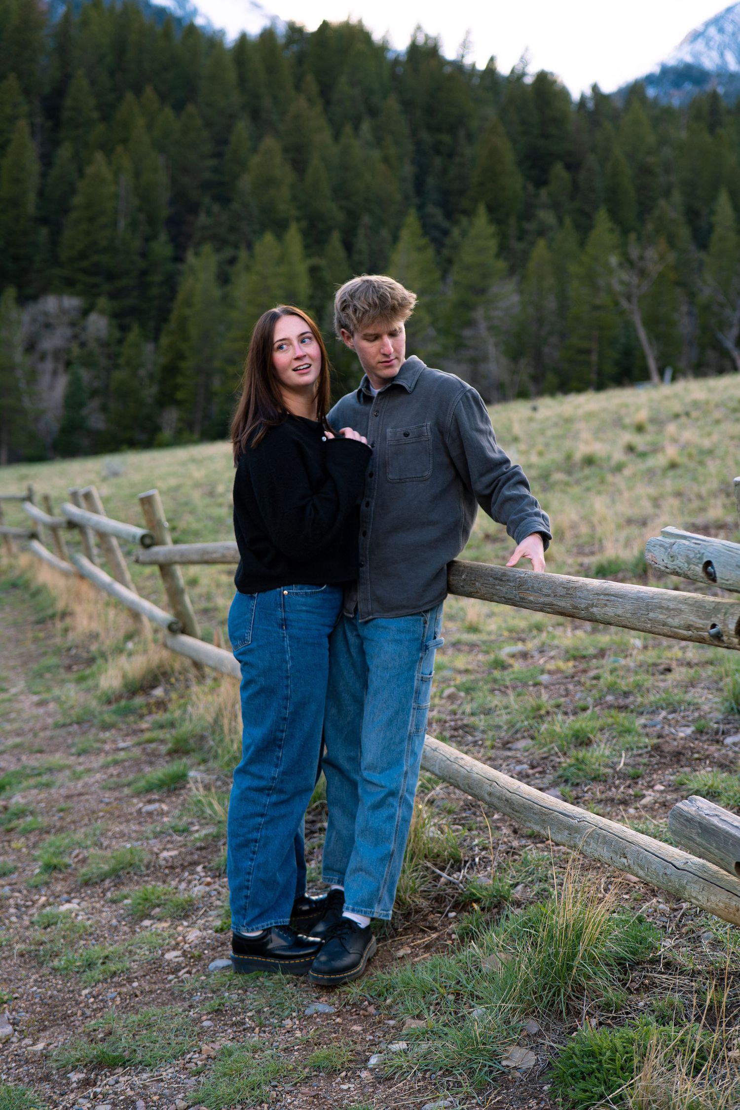 Couple posing by rustic wooden fence in mountain meadow, woman in black sweater and man in gray shirt, both wearing jeans.