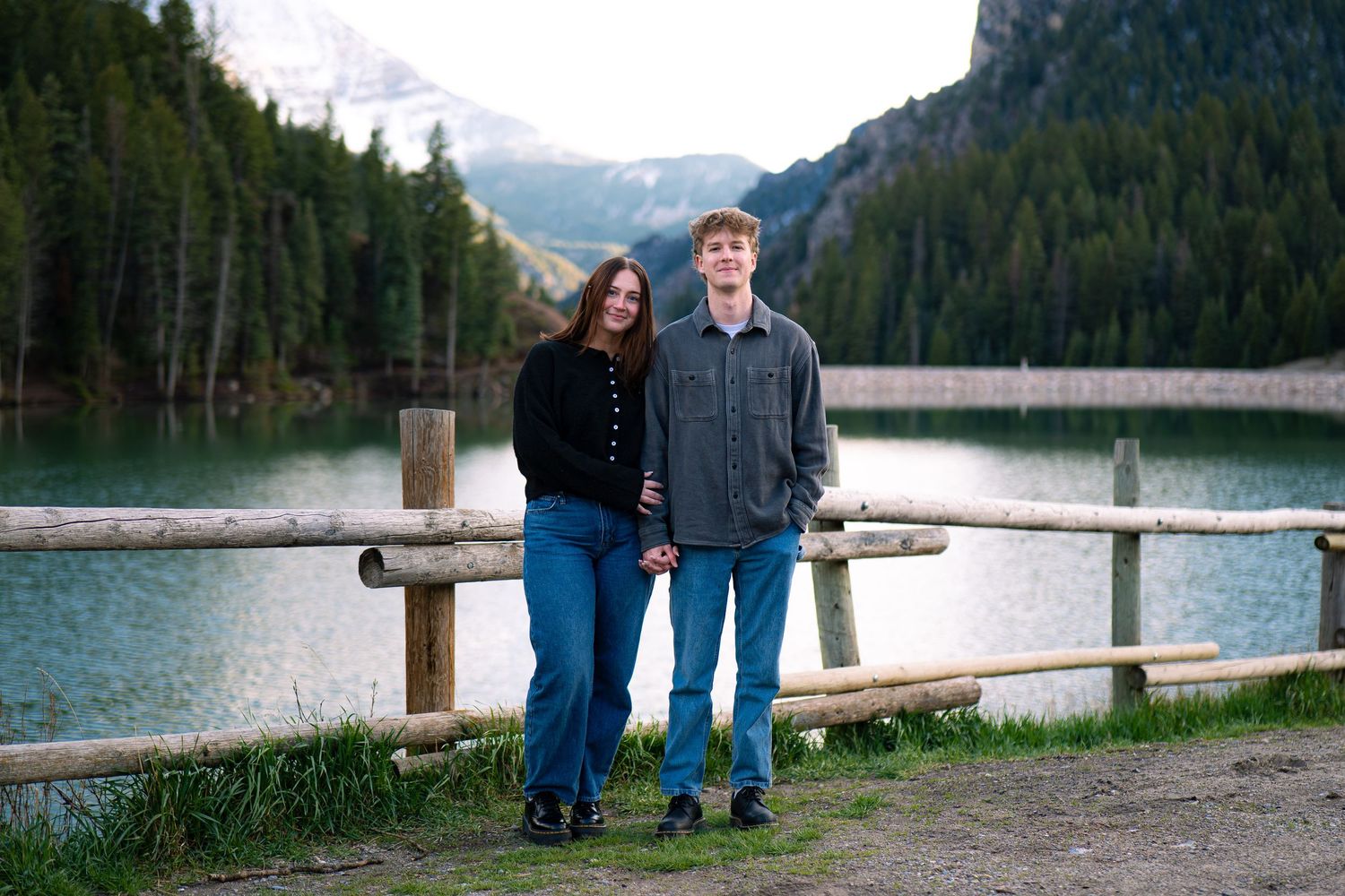 Couple holding hands by a wooden fence at a mountain lake with evergreen trees and snowy peaks.