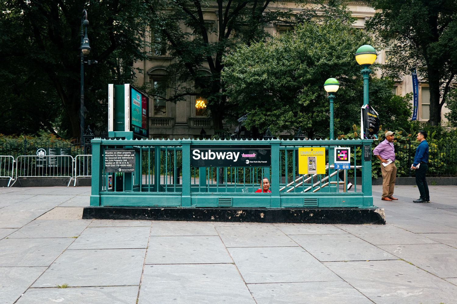 The image shows a subway entrance in New York City, with the familiar green and yellow Subway signage and a crowd of people passing through.