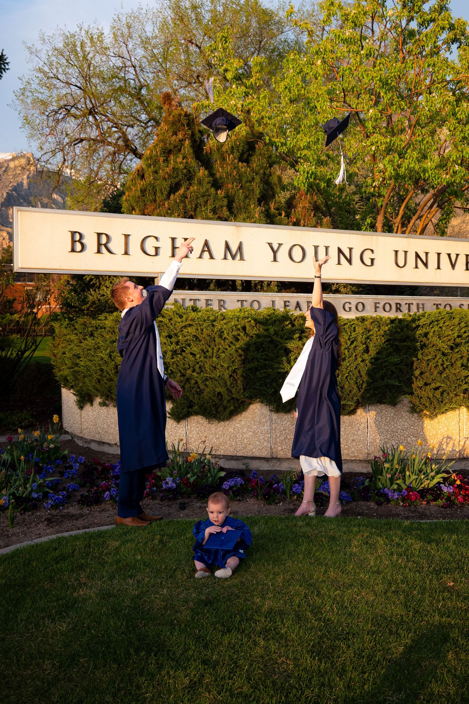 Two people in graduation gowns are standing in front of a Brigham Young University sign.