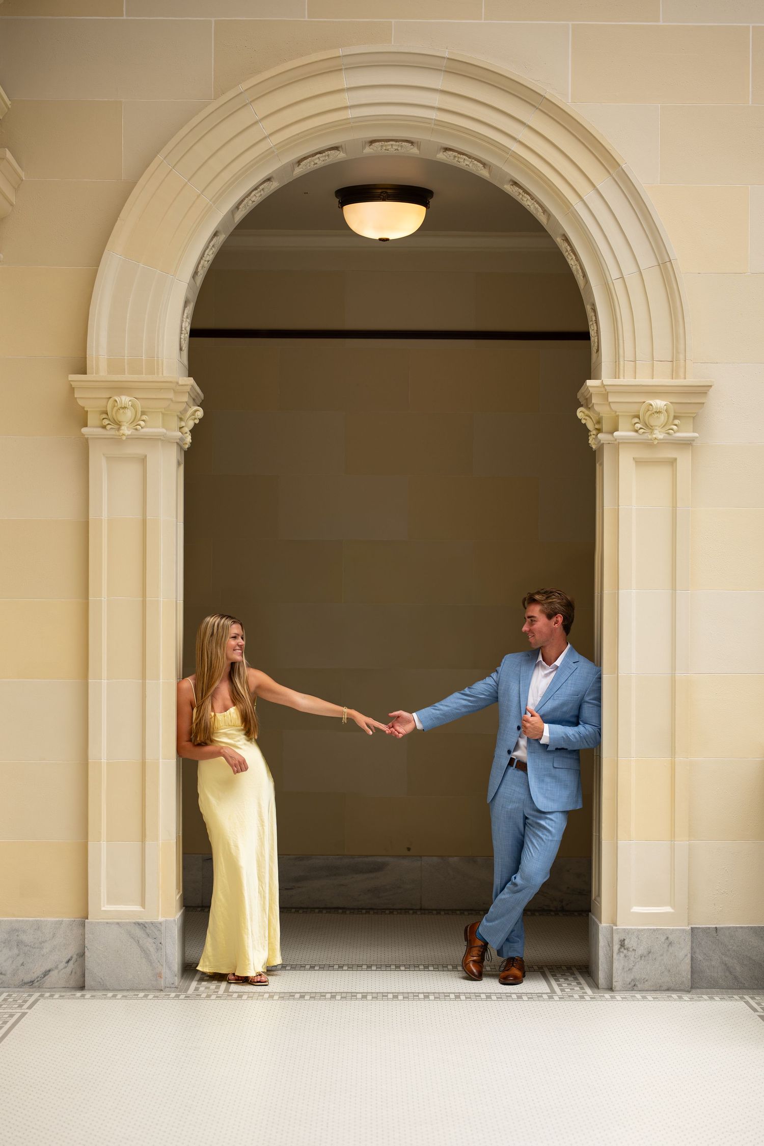 Couple holding hands under elegant stone archway, woman in yellow gown and man in blue suit.