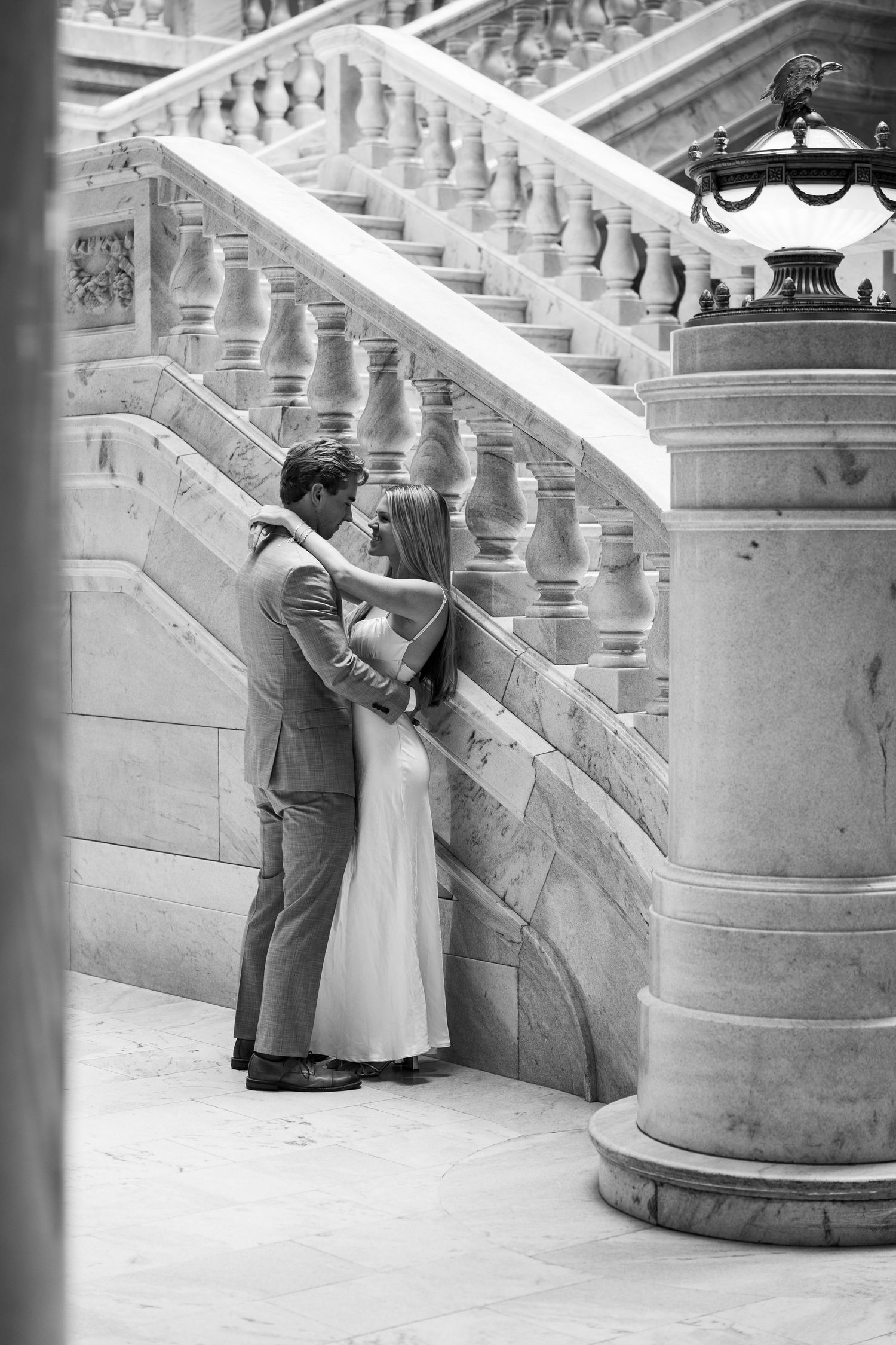 Black and white photo of a couple sharing a romantic kiss beside an elegant marble staircase with ornate balusters.