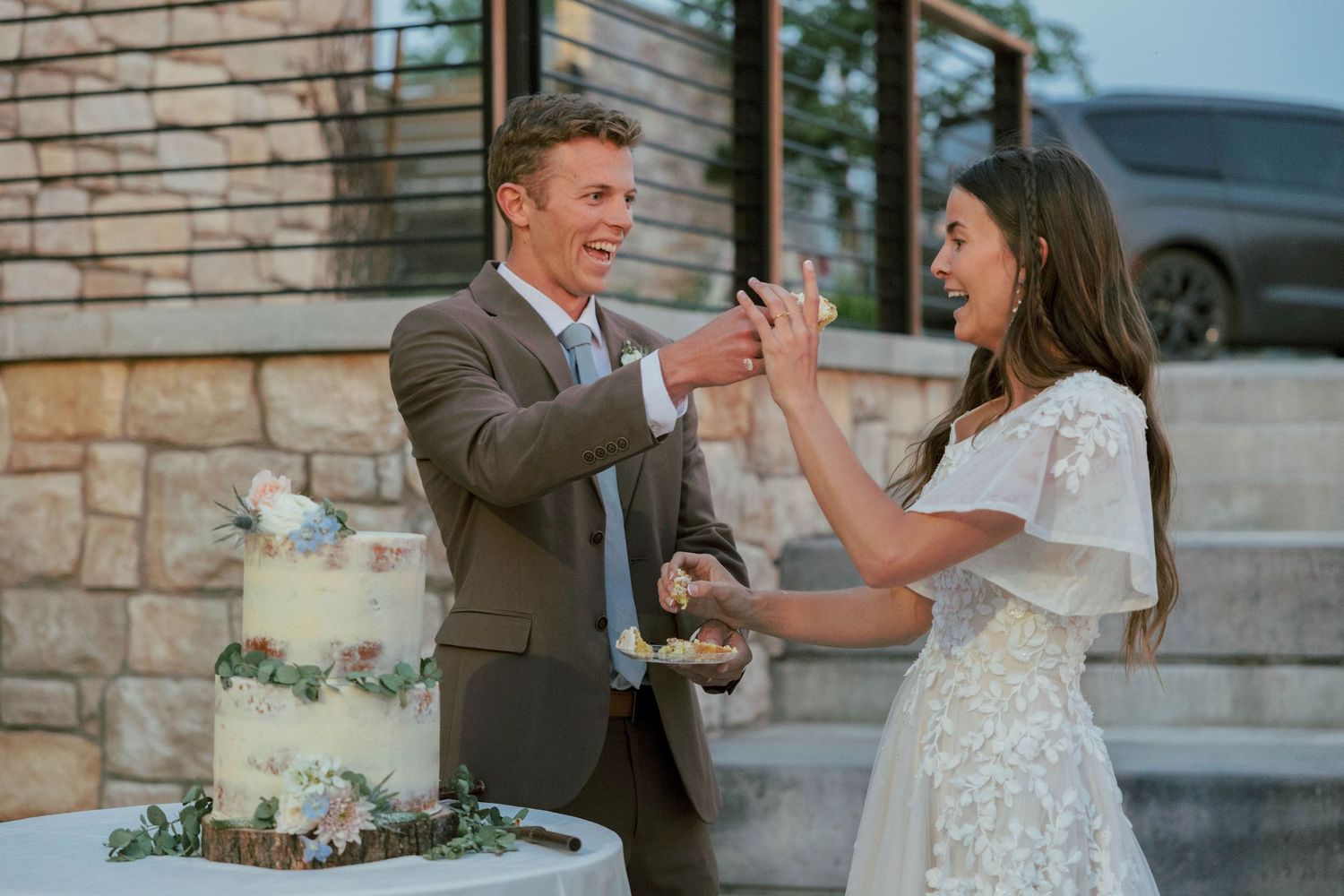 Bride and groom laughing while opening champagne bottle next to white wedding cake outdoors at sunset.