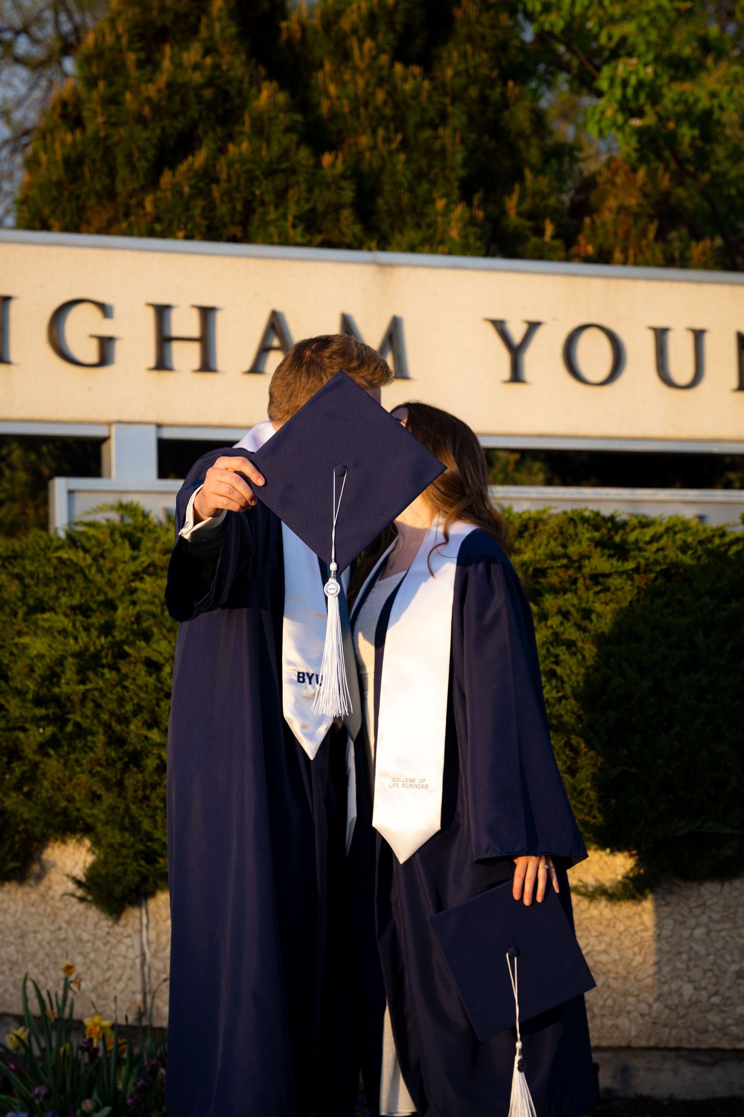 A couple in graduation gowns and caps embrace in front of a university building.