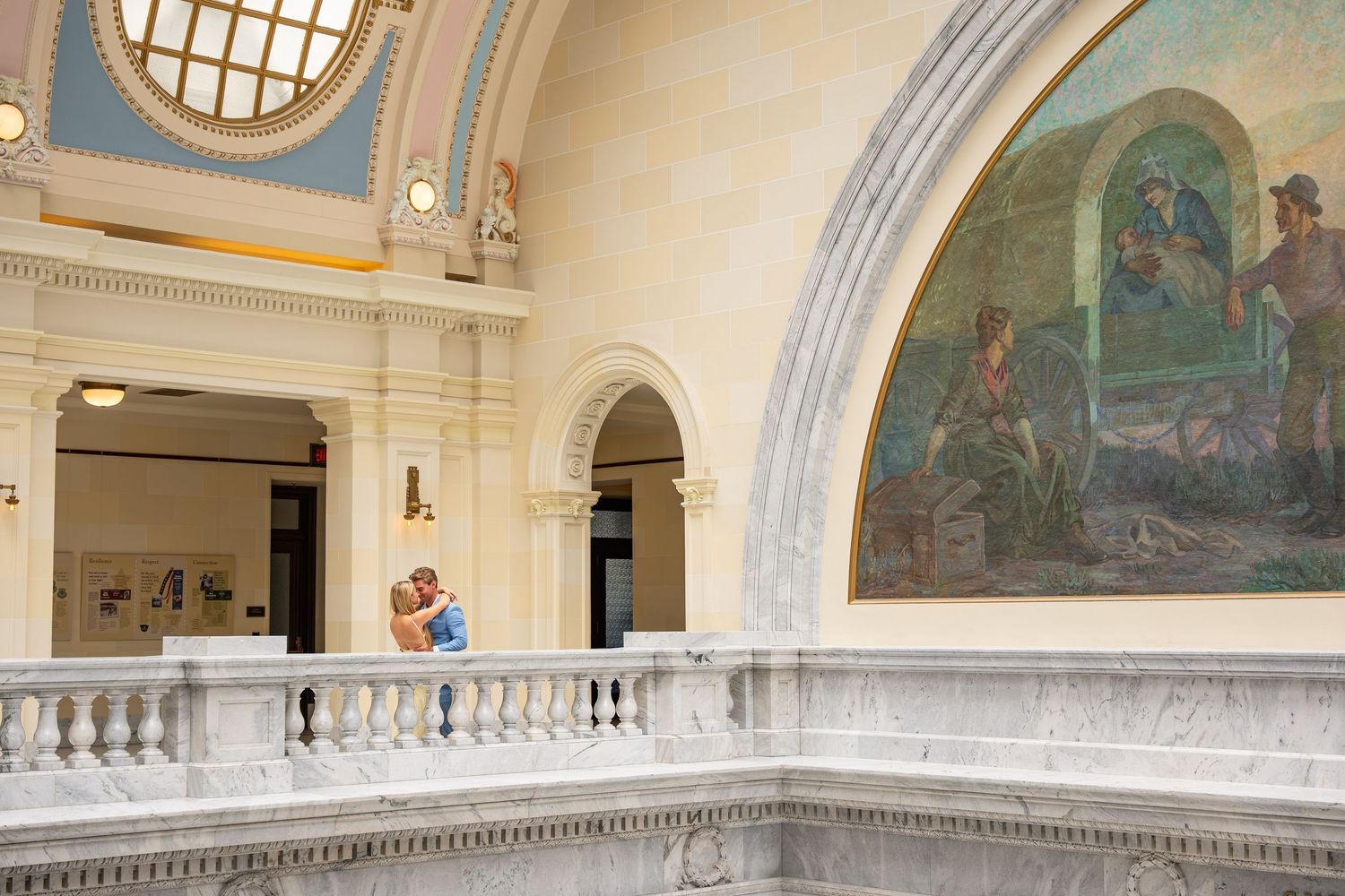 Couple embracing on marble balcony inside ornate capitol building with classical mural and domed ceiling.