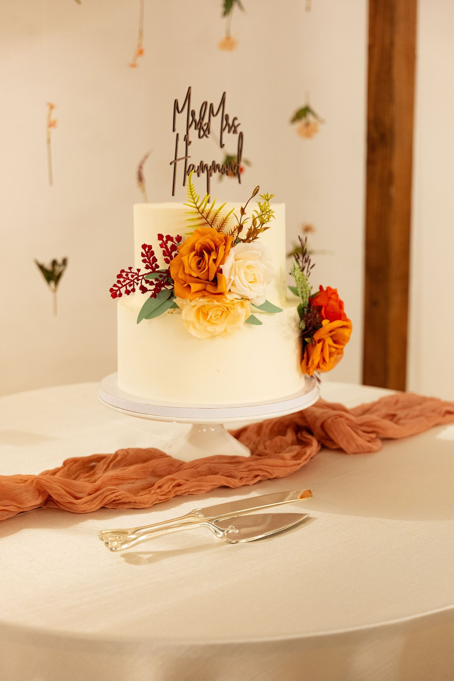 Two-tier white wedding cake with orange and yellow flowers and Mr & Mrs topper on a decorated table.