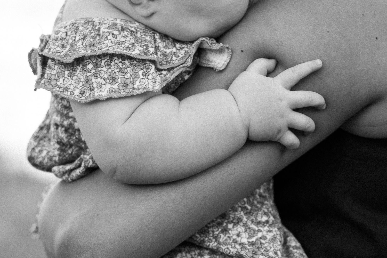 Black and white photo of a baby in a floral dress being held by an adult, showcasing tiny chubby hands.