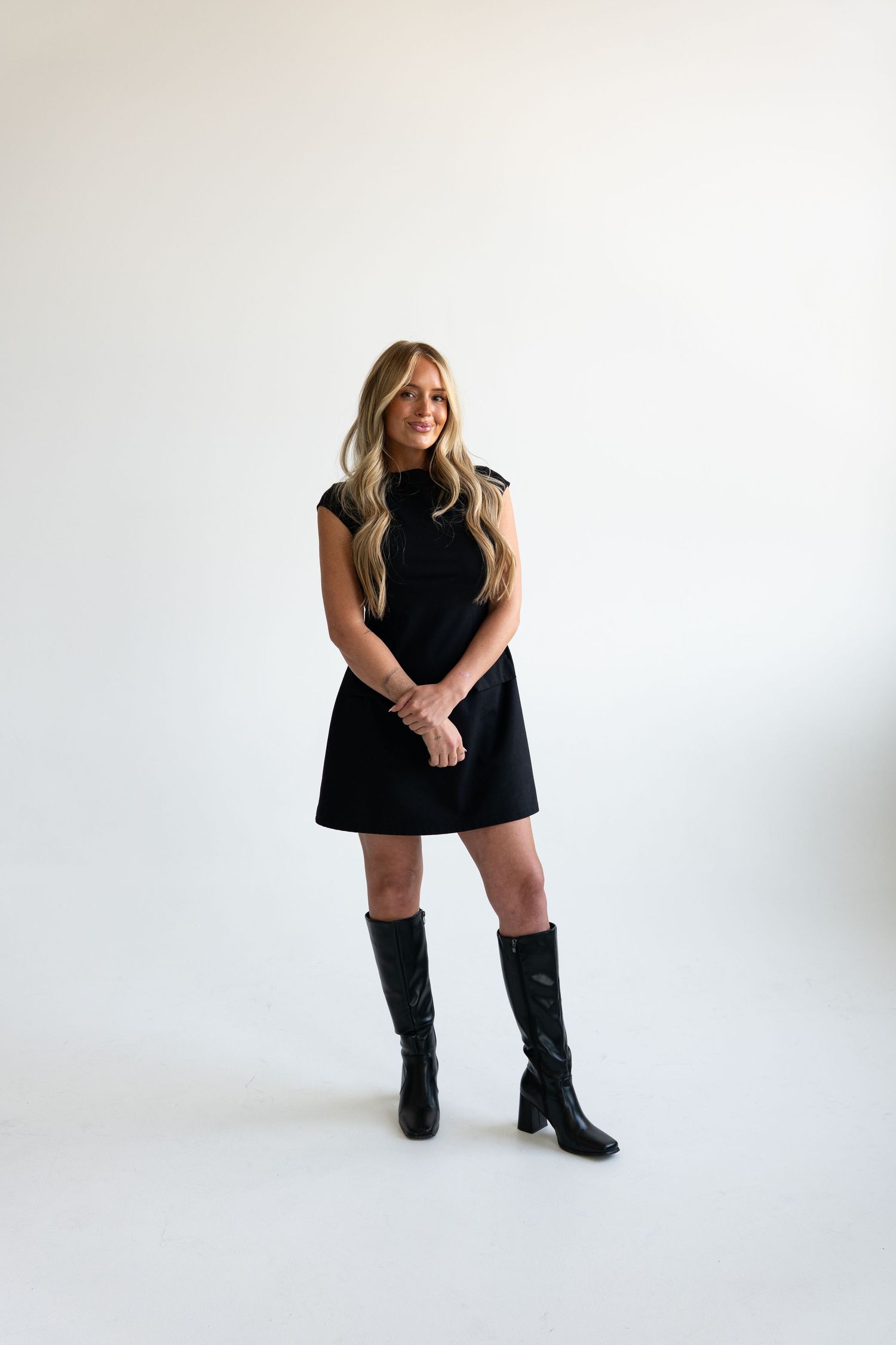 Blonde woman in black mini dress and knee-high boots smiling against white background.