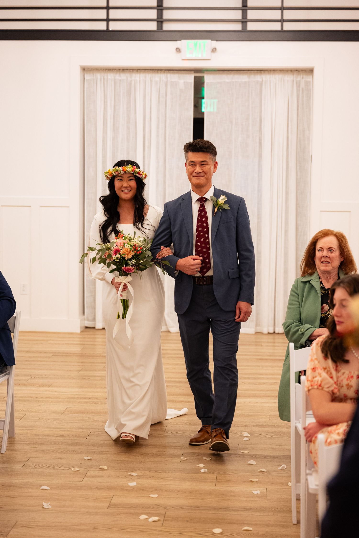 Bride in white gown with floral crown walks down aisle with father at indoor wedding.