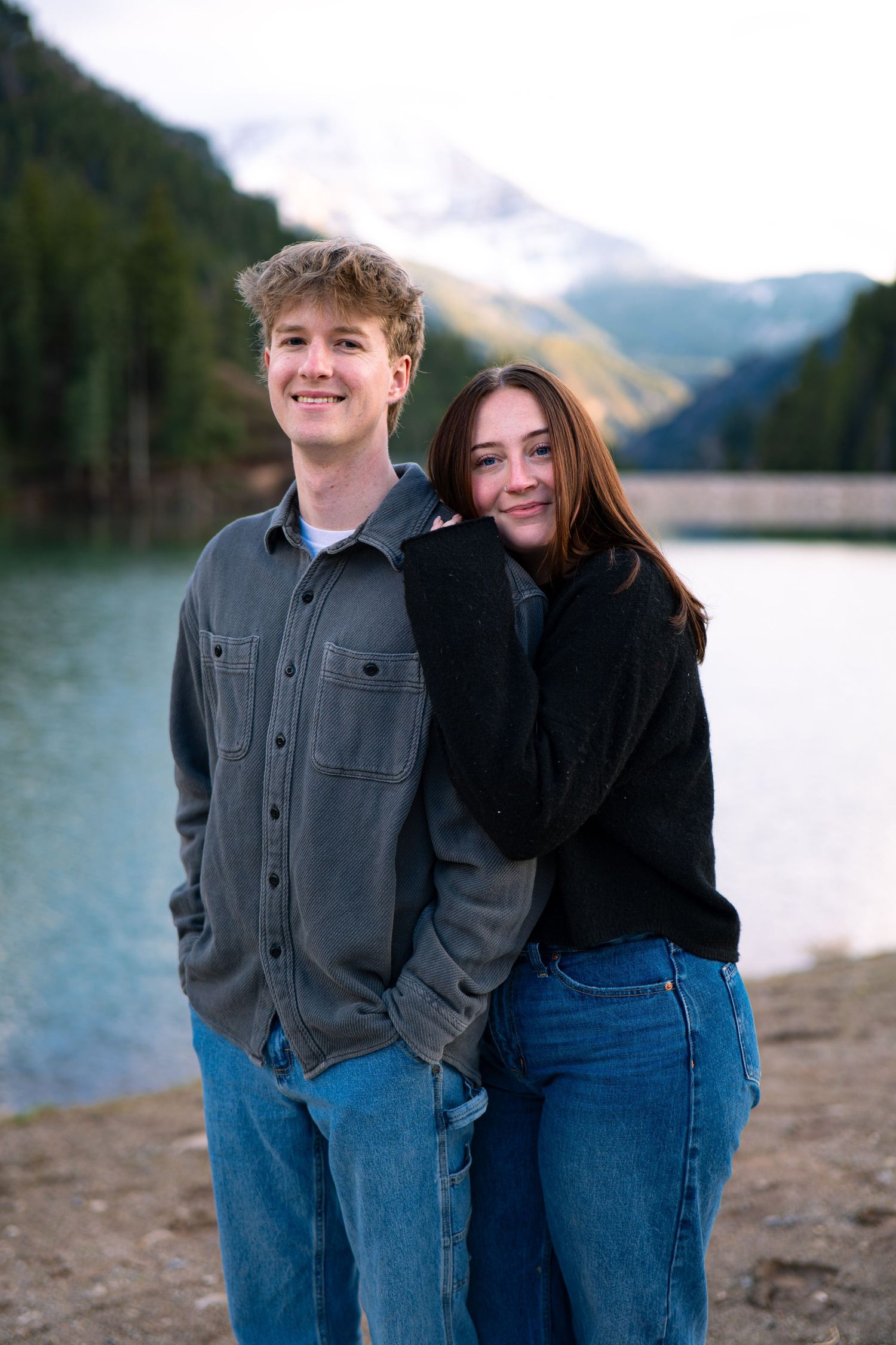 Young couple smiling by a mountain lake, wearing casual jeans and jackets, with snow-capped peaks in the background.