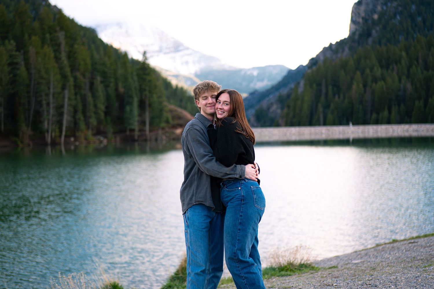Couple embracing by a mountain lake with snow-capped peaks and pine forest backdrop.