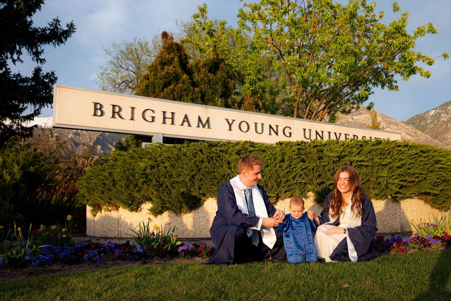 A family of three, including a young child, stands in front of a sign for Brigham Young University.