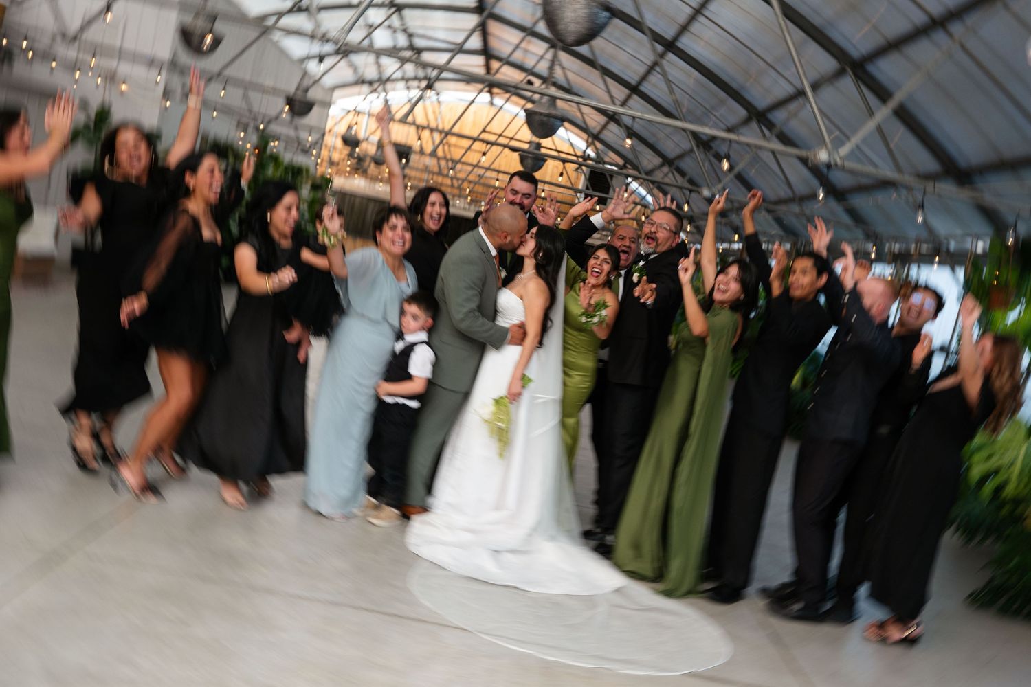 Wedding party celebrating with bride and groom under elegant tent venue, bridesmaids in green dresses and groomsmen in black suits.