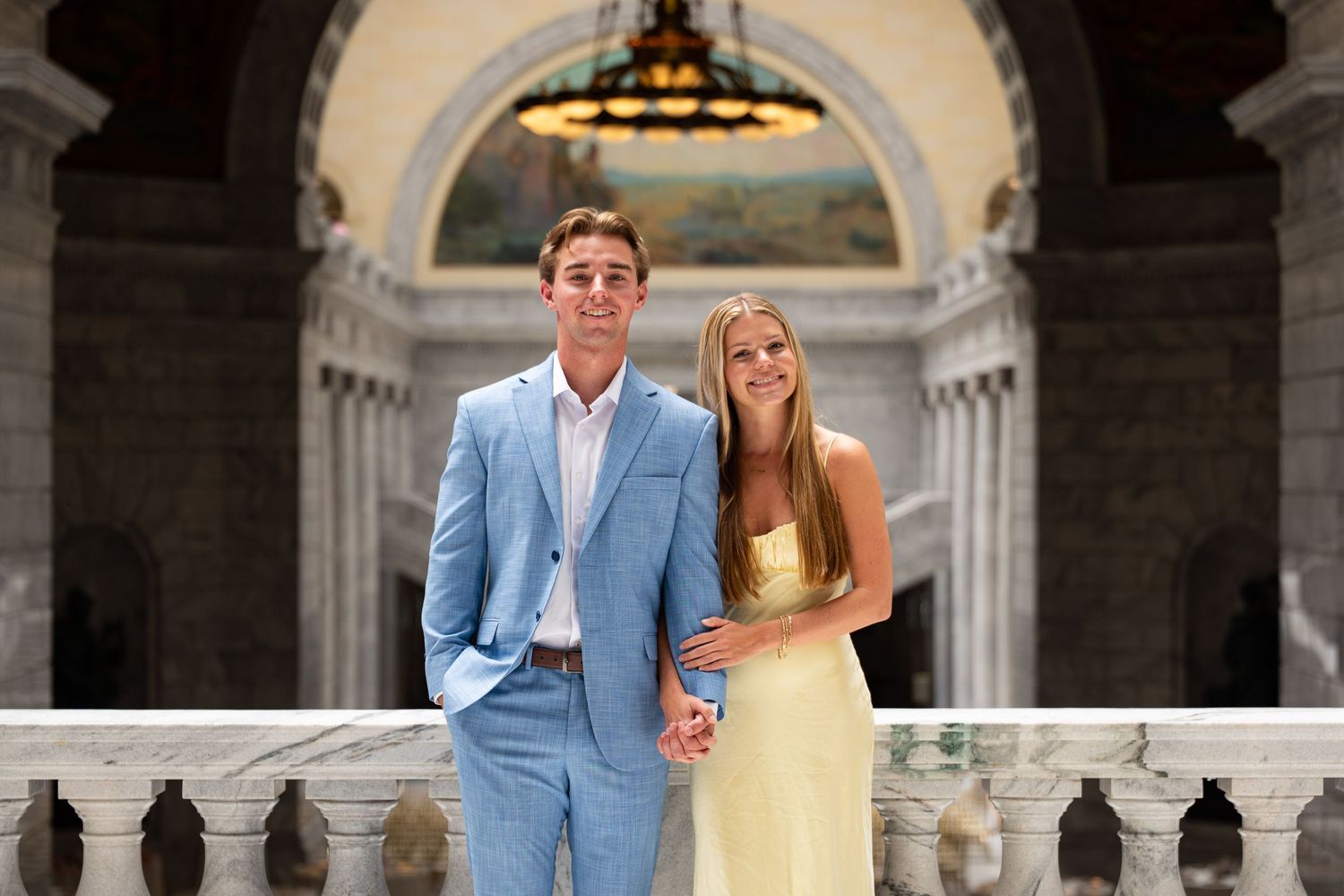 Young couple in formal attire posing in elegant marble building, man in blue suit, woman in yellow dress.