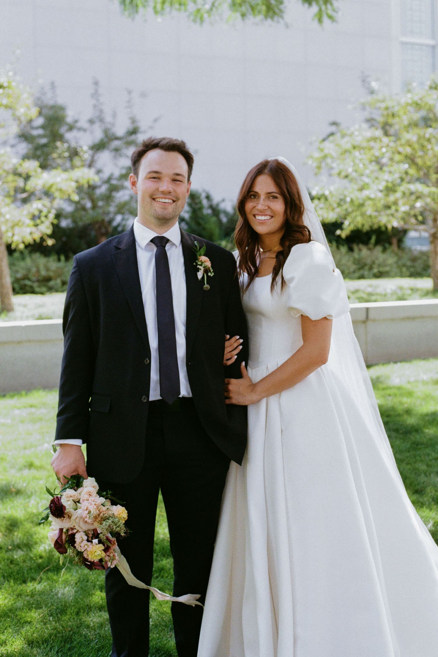 A couple in formal attire, a bride in a white dress and a groom in a black suit, stand together and smile.