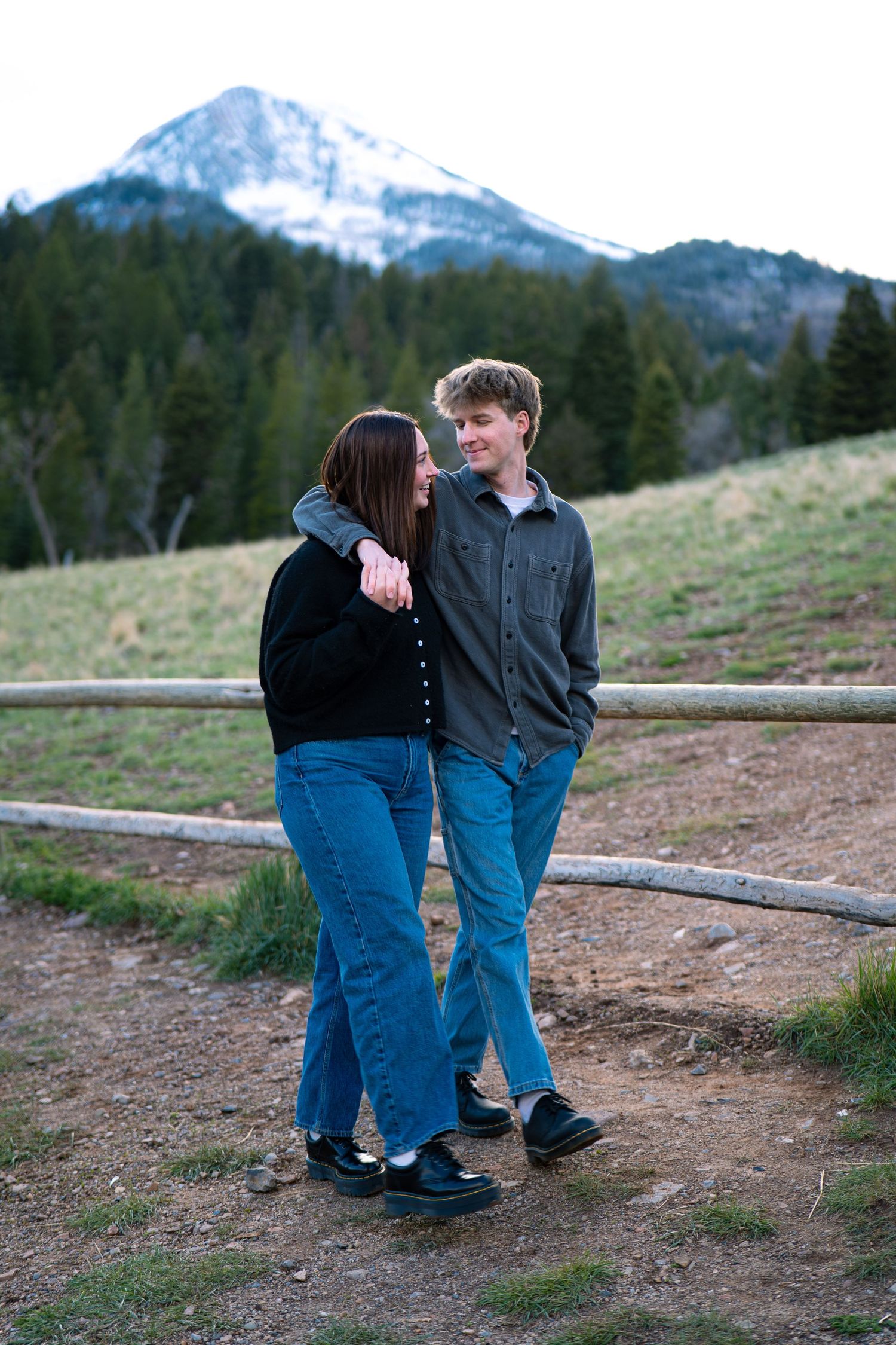 Young couple embracing and smiling at each other outdoors with a snow-capped mountain and pine trees in the background.