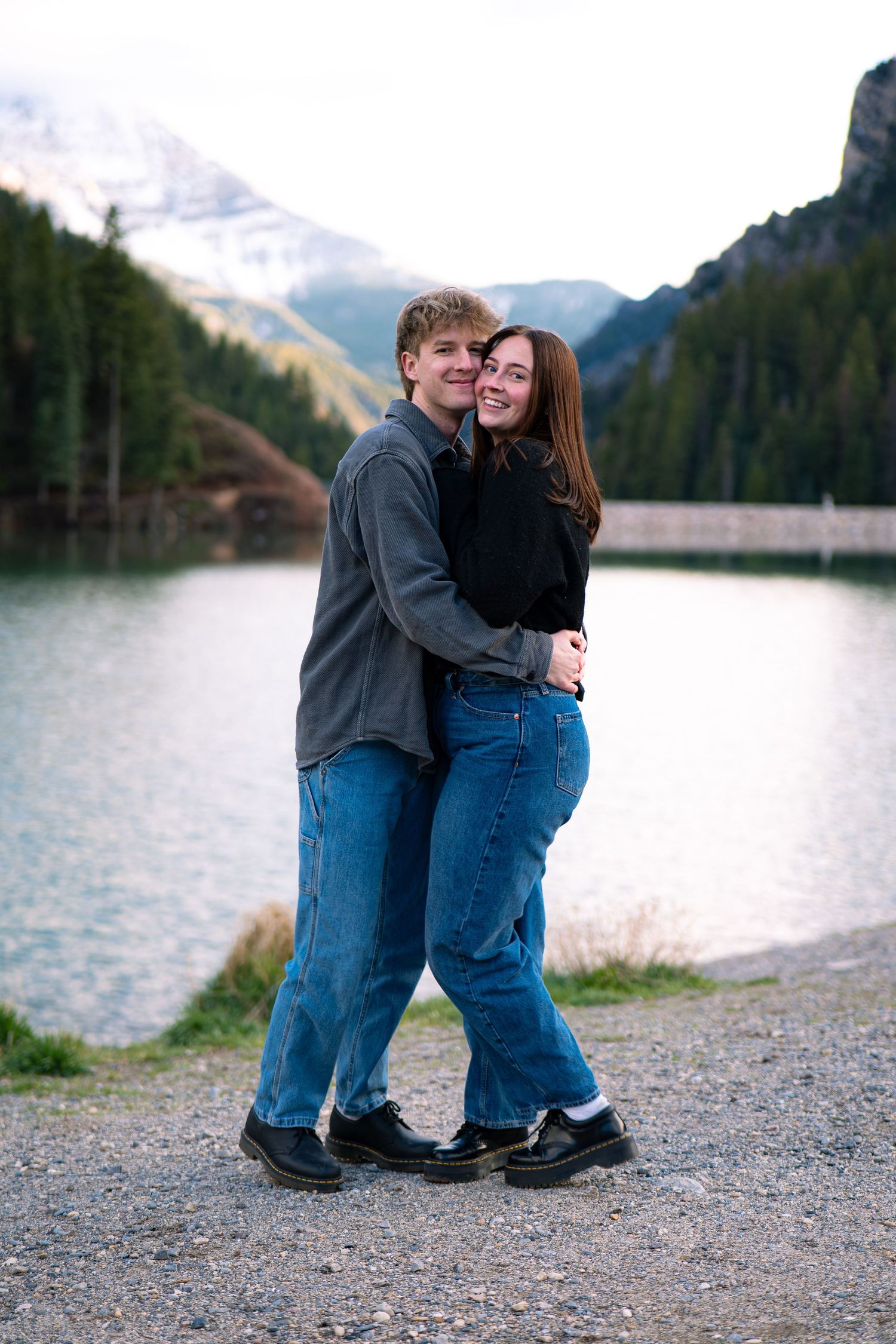 A smiling couple embraces by a mountain lake with snow-capped peaks and pine trees in the background.