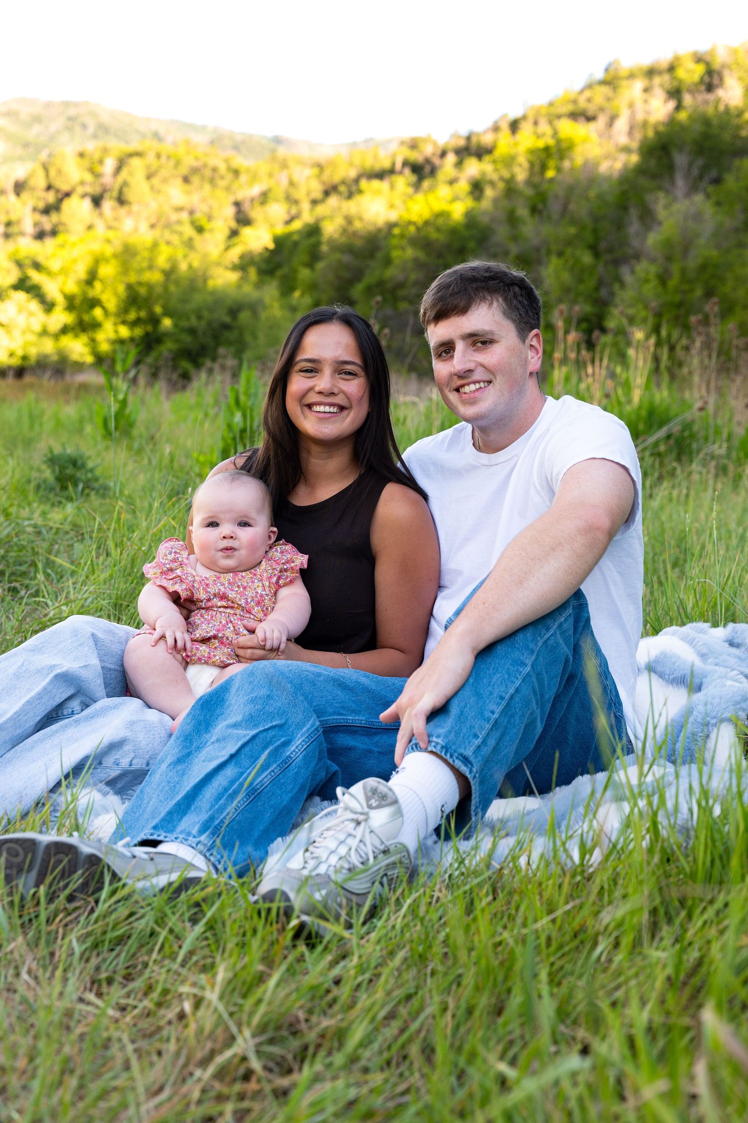 Young family with baby sitting on blanket in green meadow with tree-covered hills in background.