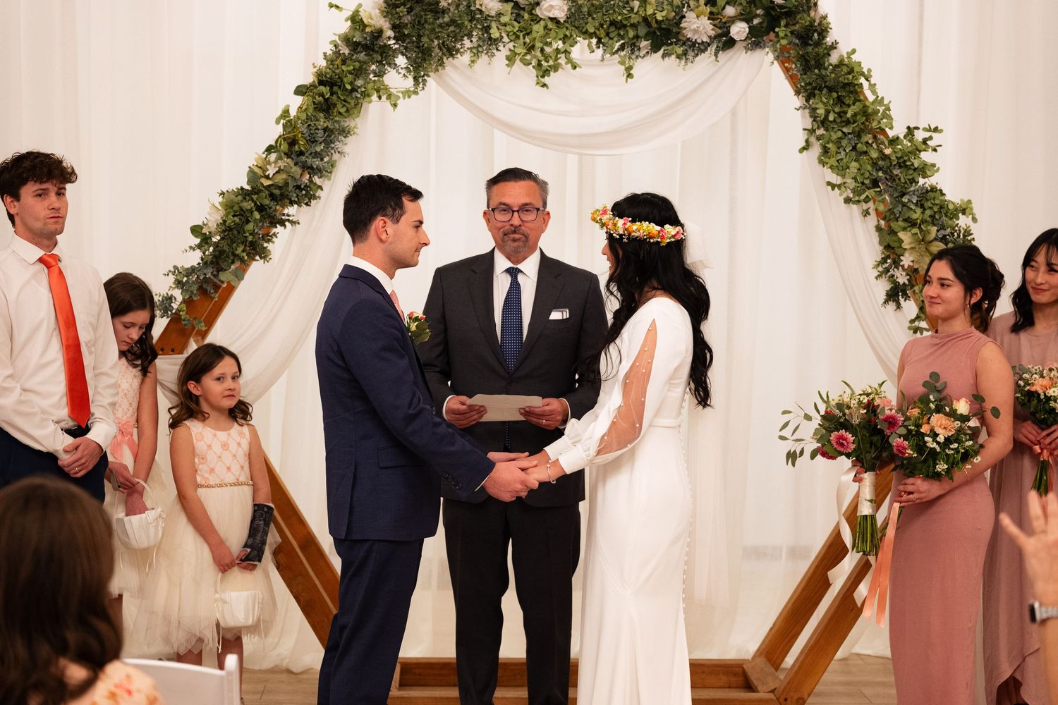 Bride and groom exchange vows at altar with greenery arch and wedding party.