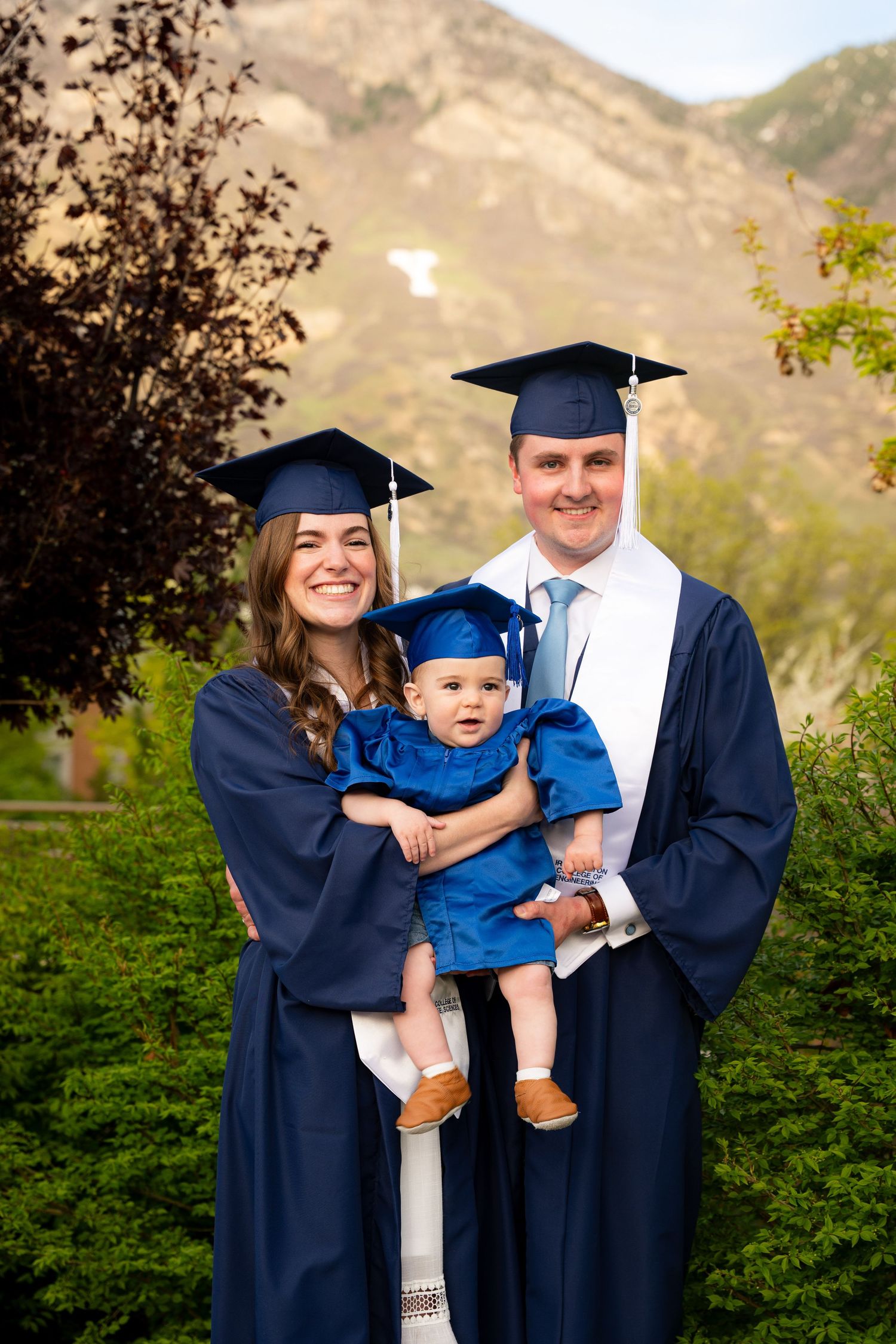 A family of three, including a baby, pose for a graduation photo outdoors.