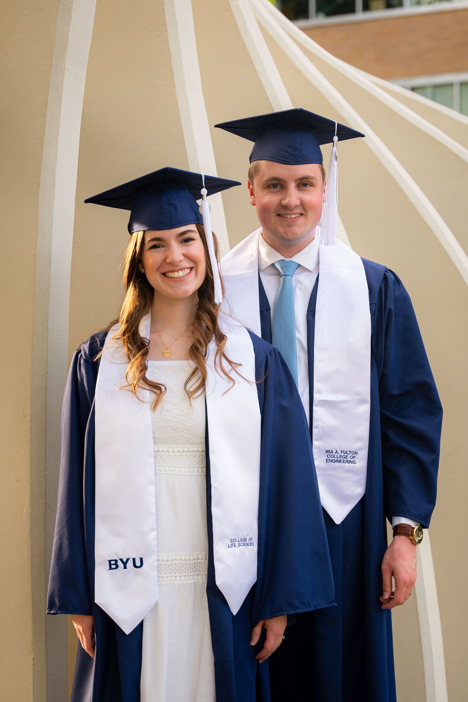 A couple in graduation gowns and caps stand together in front of a university building.