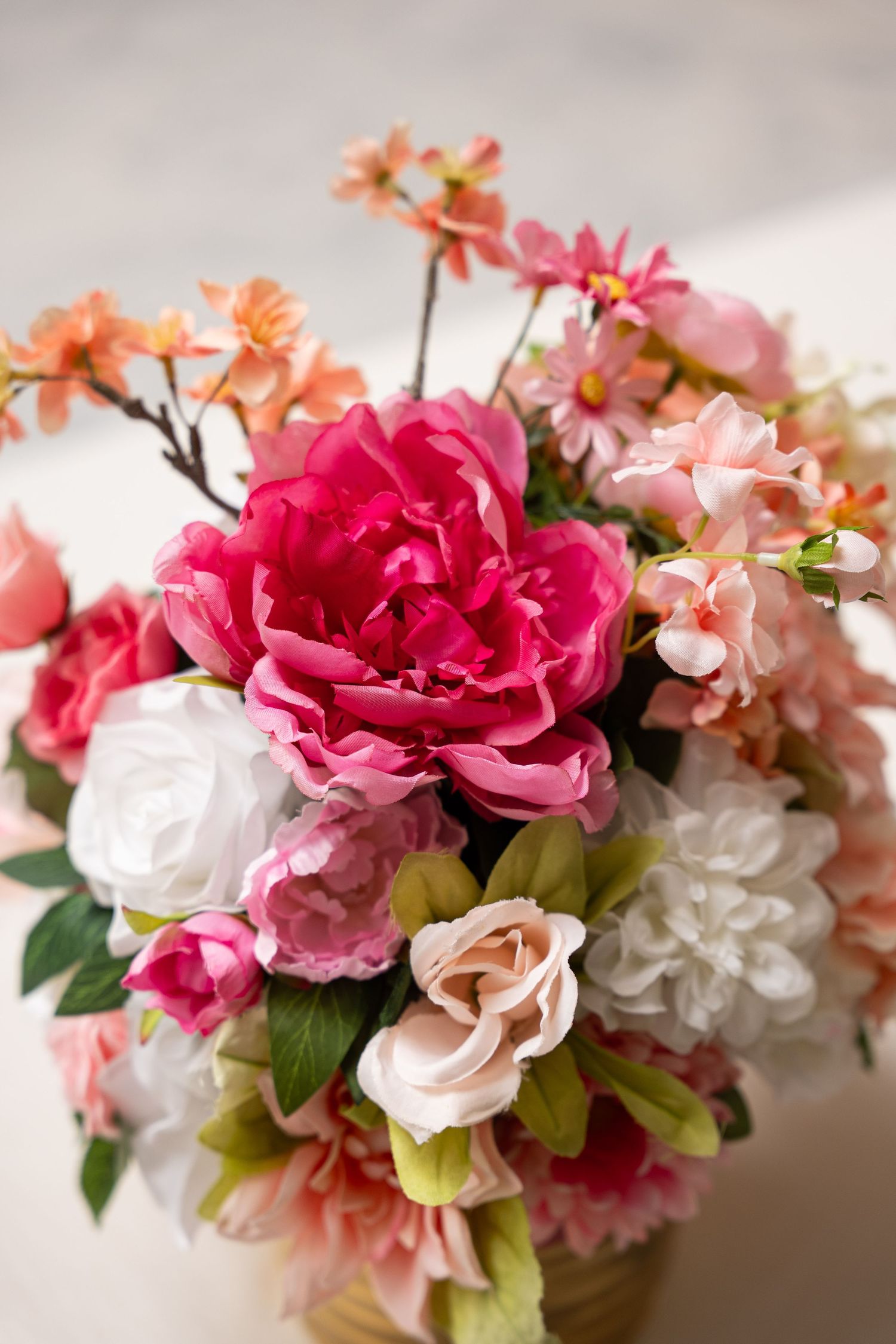 Vibrant bouquet of pink peonies, white roses, and coral blossoms arranged in a vase on a light background.