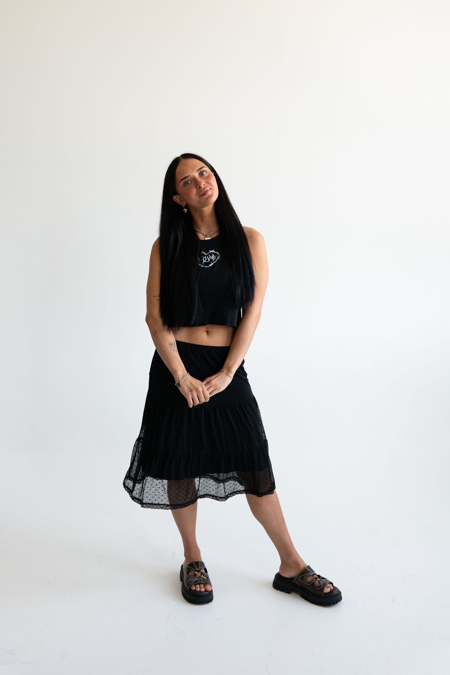Woman in black crop top, midi skirt, and sandals posing against white background.