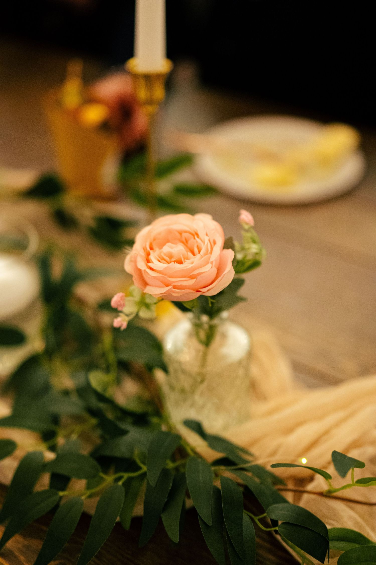 Peach garden rose in glass vase with eucalyptus greenery on rustic wooden wedding reception table.