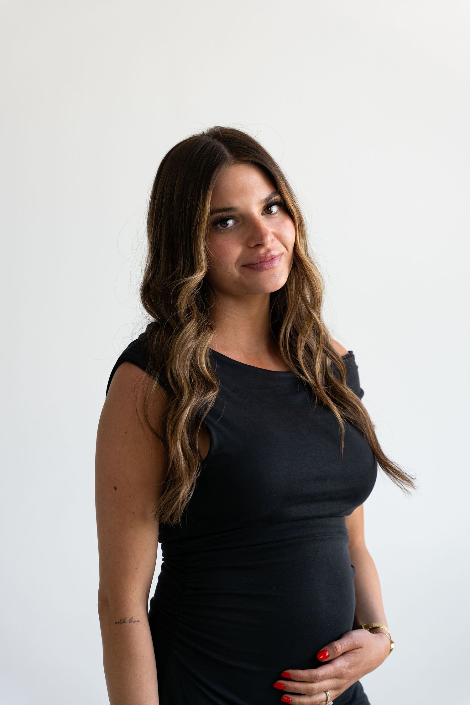 Pregnant woman in black dress cradling baby bump, smiling softly with long wavy hair against white background.