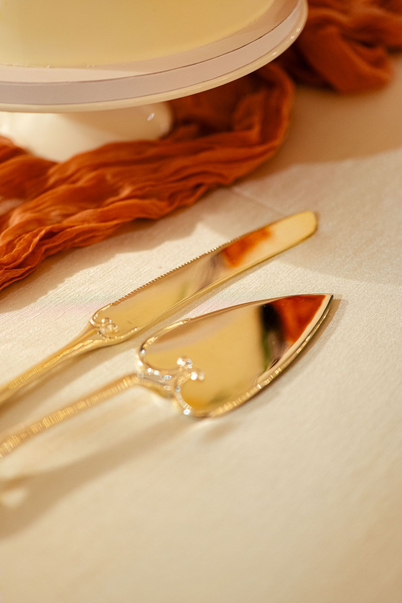 Gold cake knife and server set on white linen tablecloth with rust orange ribbon and white cake stand.