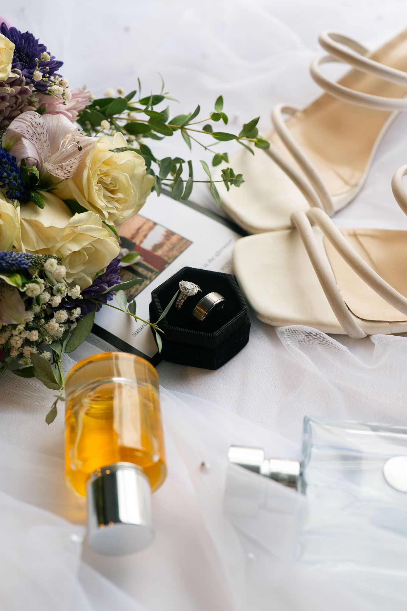 Bridal flat lay with bouquet, cream heels, wedding rings in velvet box, and perfume bottles on white fabric.